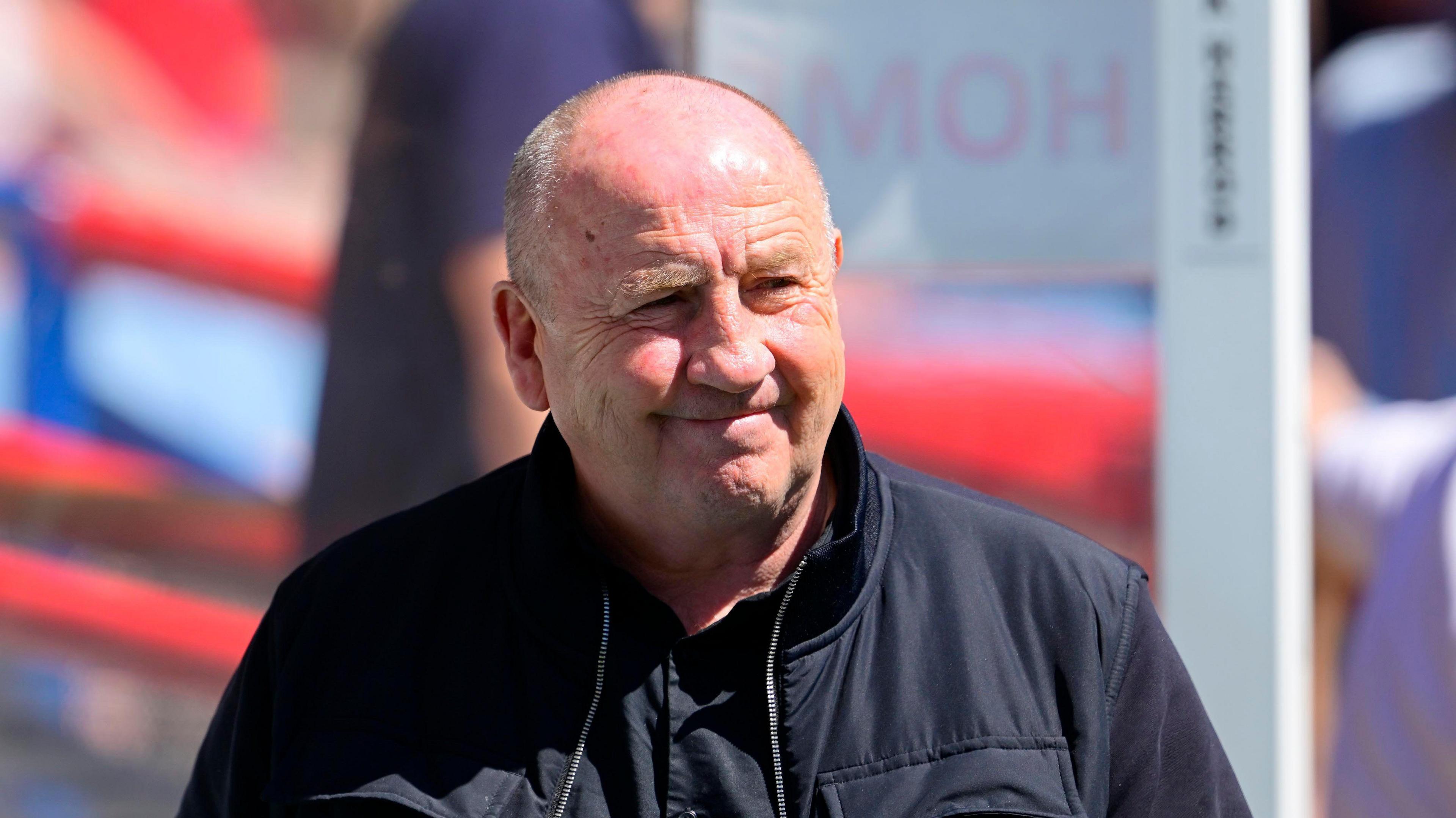 John Coleman smiling next to the dugout, looking across the pitch in a dark shirt and matching dark jacket 