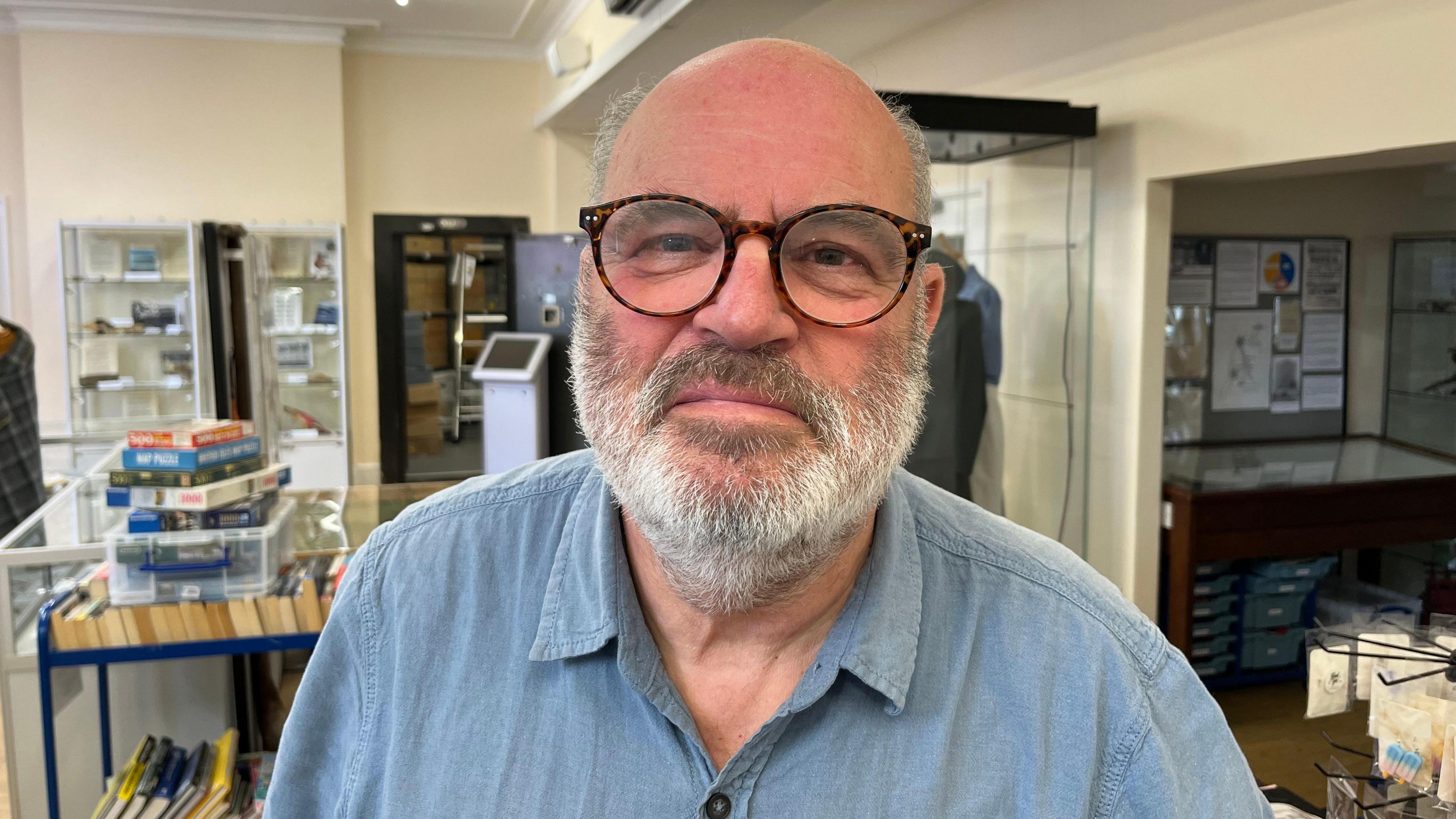 A bald man with thin grey hair on the side of his head. He has a grey beard and moustache and is wearing brown glasses. He is wearing a blue denim-type shirt. Behind him are items on display at the museum.