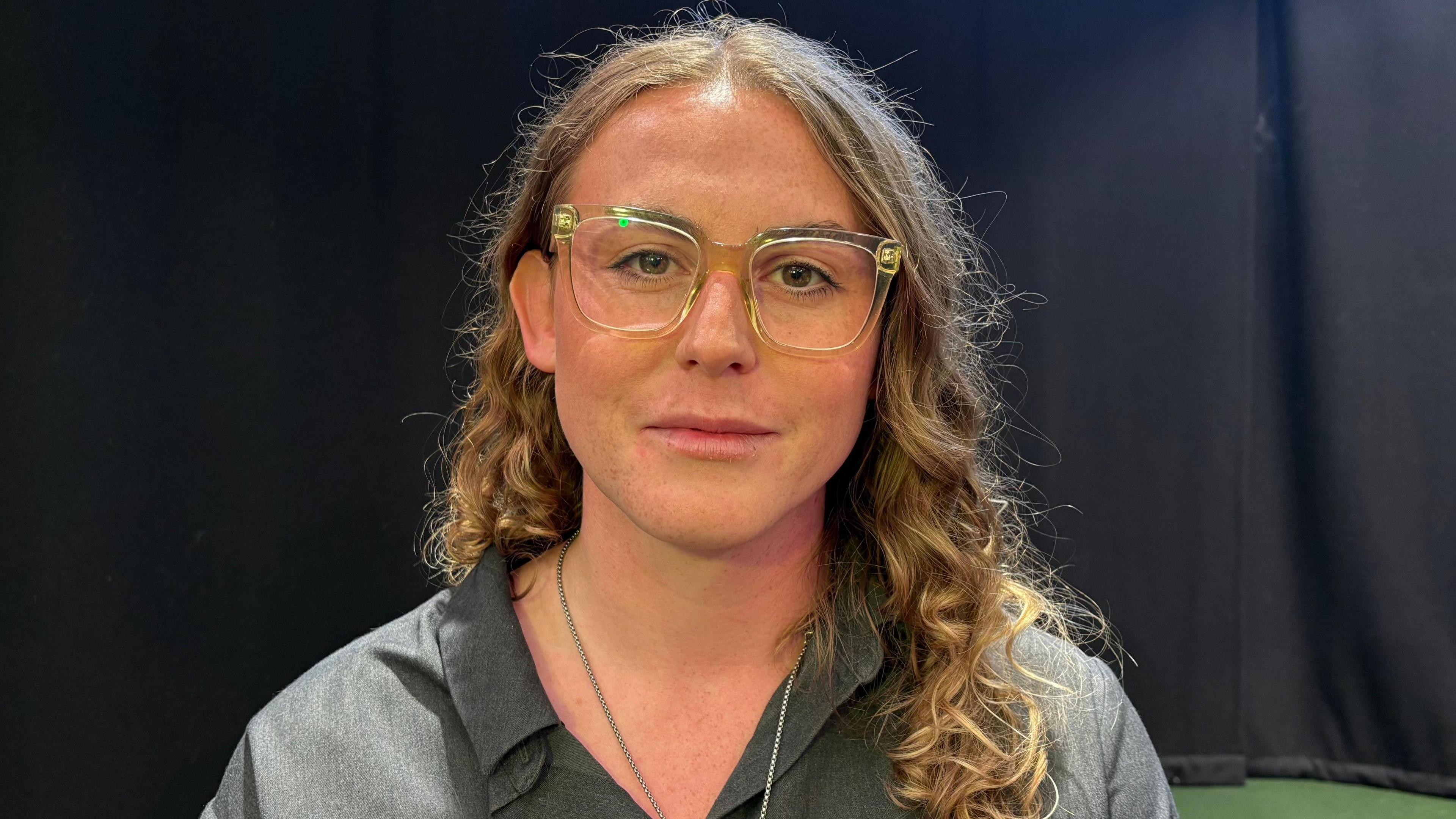Kate Lankester looking directly down the camera. She is wearing a grey, open-necked, collared top, with a silver chain around her neck, though the pendant is not in the frame. She is wearing square-framed glasses with transparent plastic frames. She has curly, light brown hair that is falling below one of her shoulders. She is sat against a black backdrop.