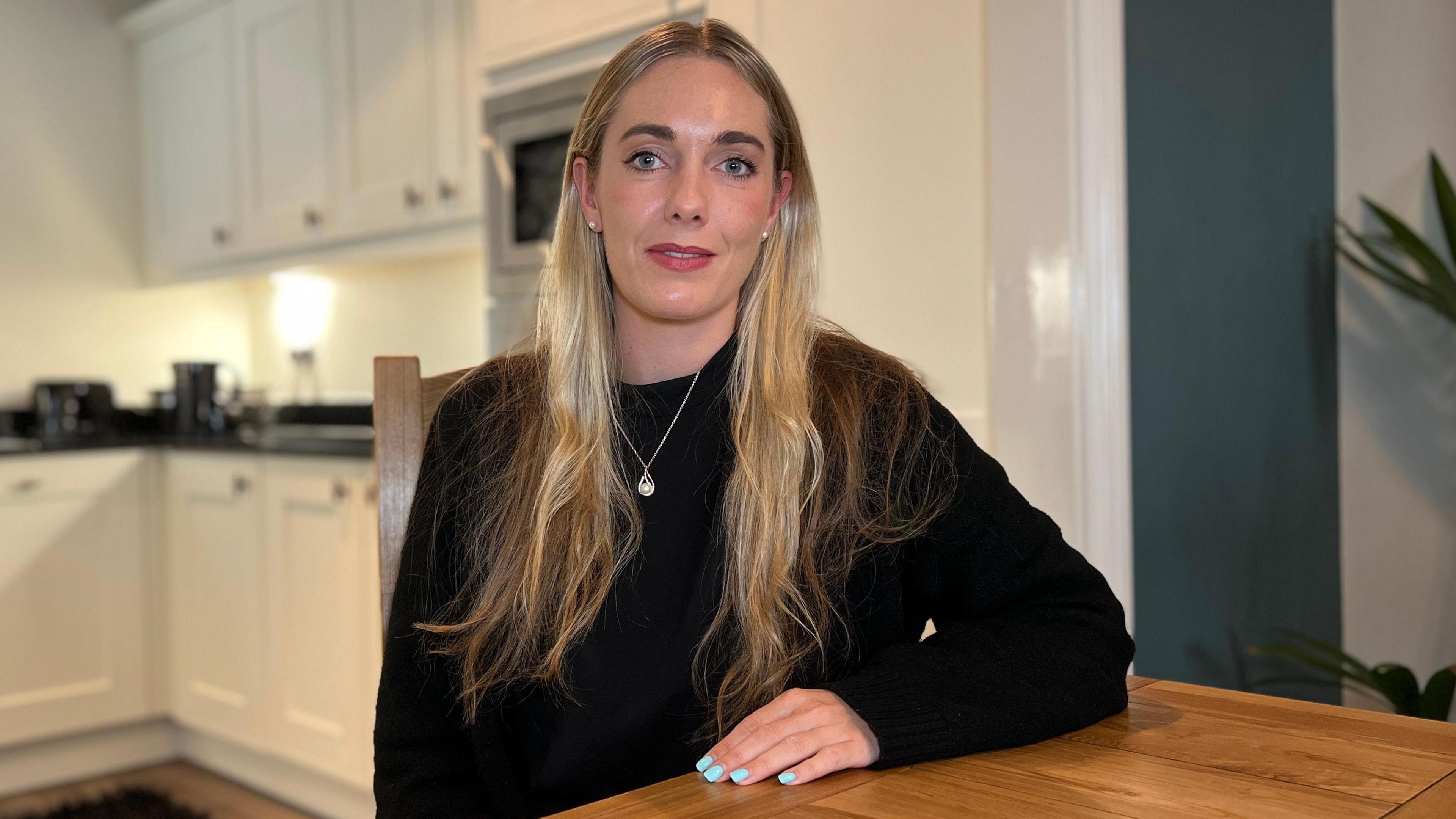 Jessica looks at the camera as she sits at a wooden table. She has a black jumper on with a silver necklace. She has long dark blonde hair. There is a kitchen in the background.