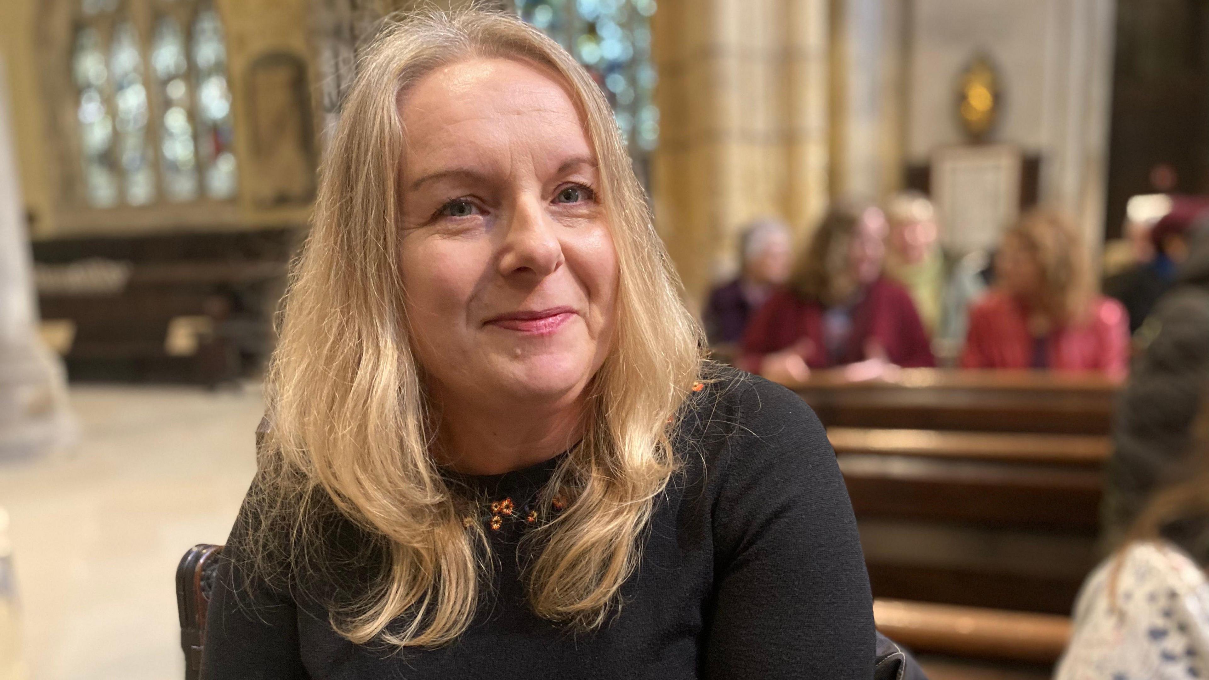 Fiona has long blond hair and a black top on. She smiles for the camera. Other people are sitting in pews behind her.