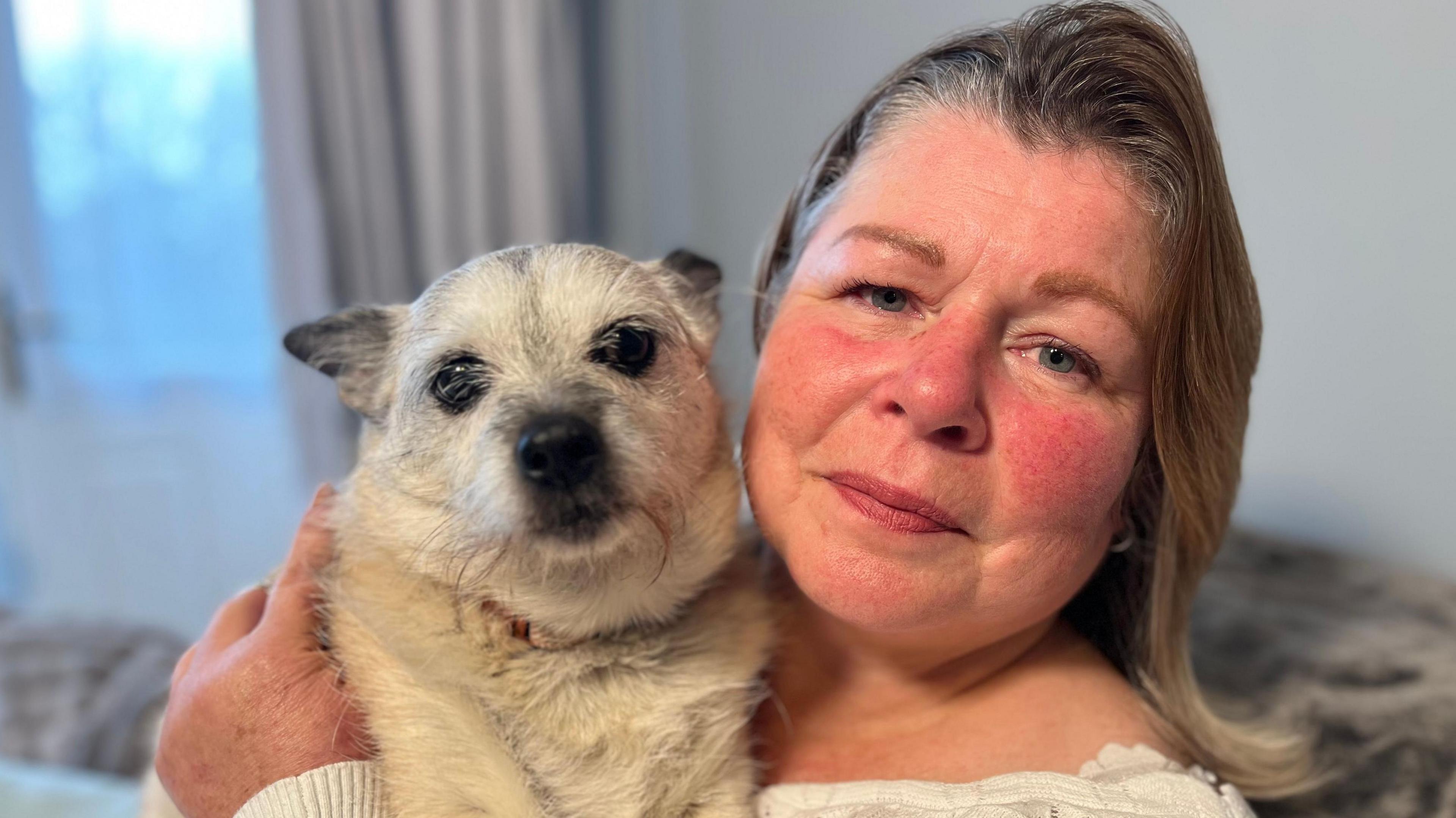 Teresa holding Morgan's white small dog Peaches. Teresa has shoulder-length fair hair. It is a head and shoulders shot.