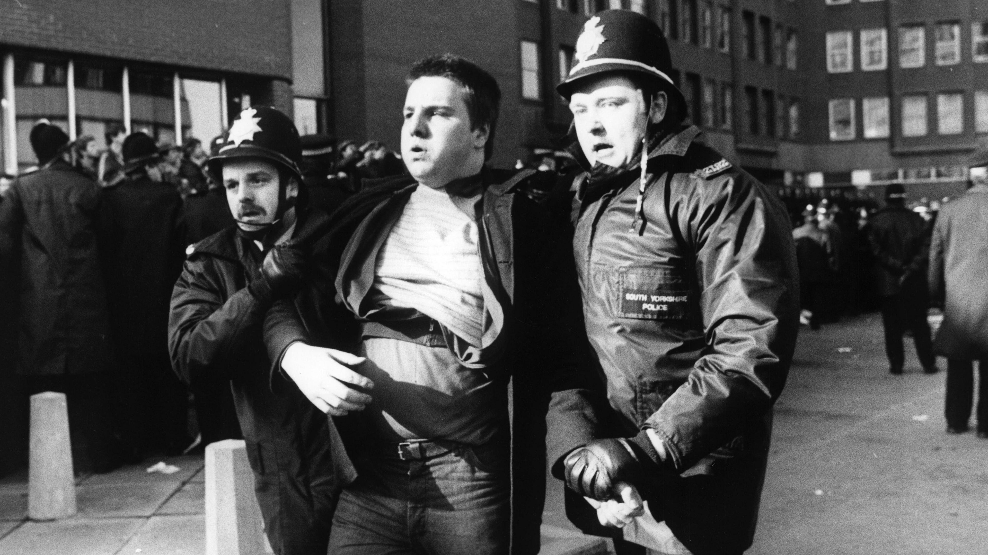 A man is led away by two police officers wearing helmets, against the backdrop of what appears to be a commotion.