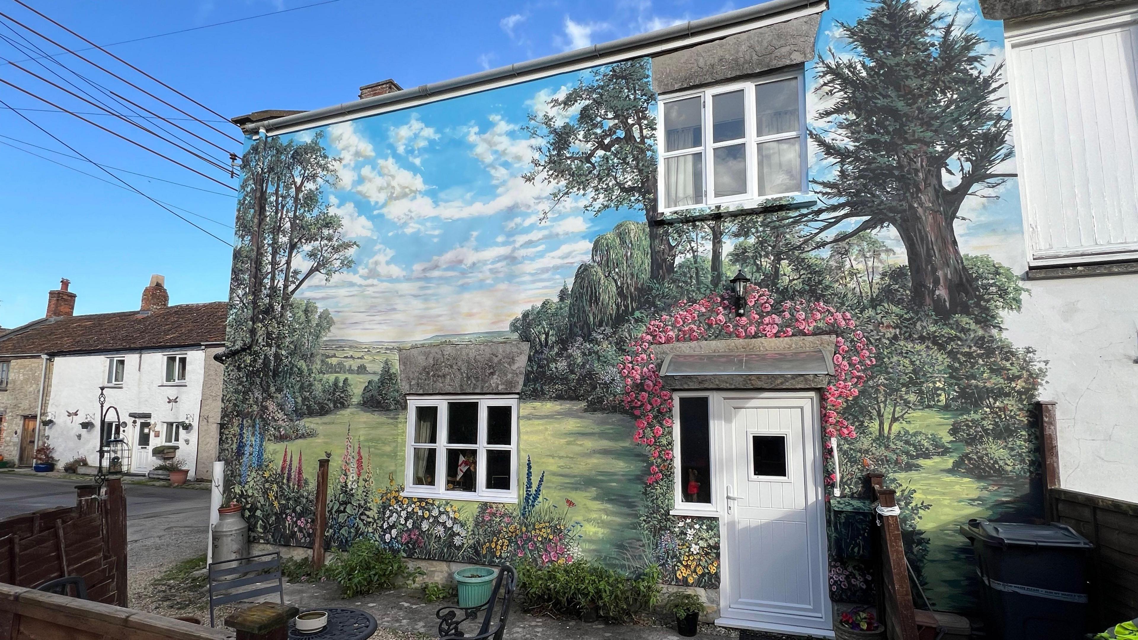 Huge mural painted on a house. She is stood near to the house's white door. The wall is painted with grass, lots of colourful flowers, butterflies and there is a countryside landscape.