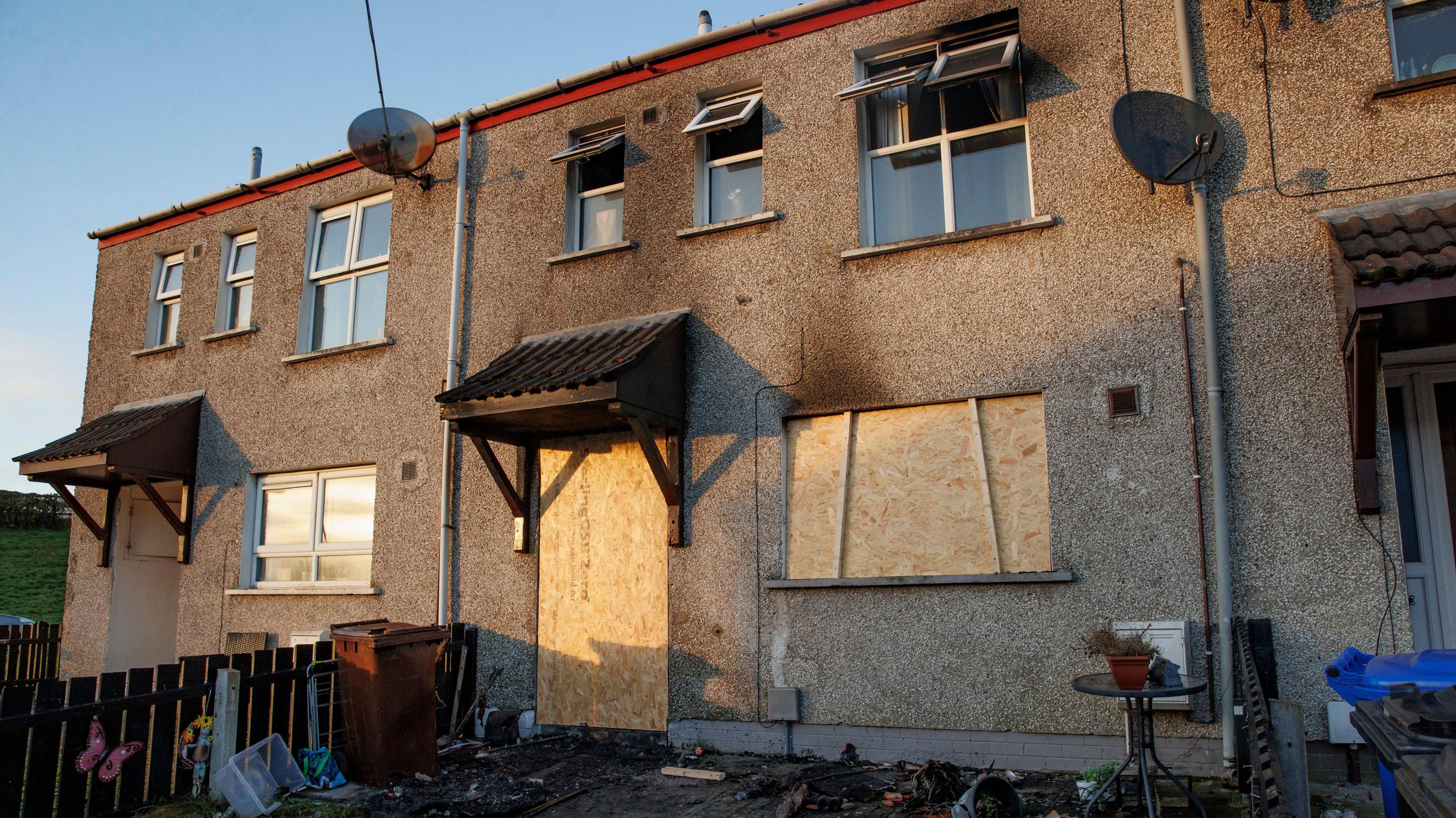 The middle house of the three was damaged in the arson. The door and downstairs window have been boarded up. The upstairs windows are open wide.  There is black marks around the windows. There is also signs of damage infront of the house. 