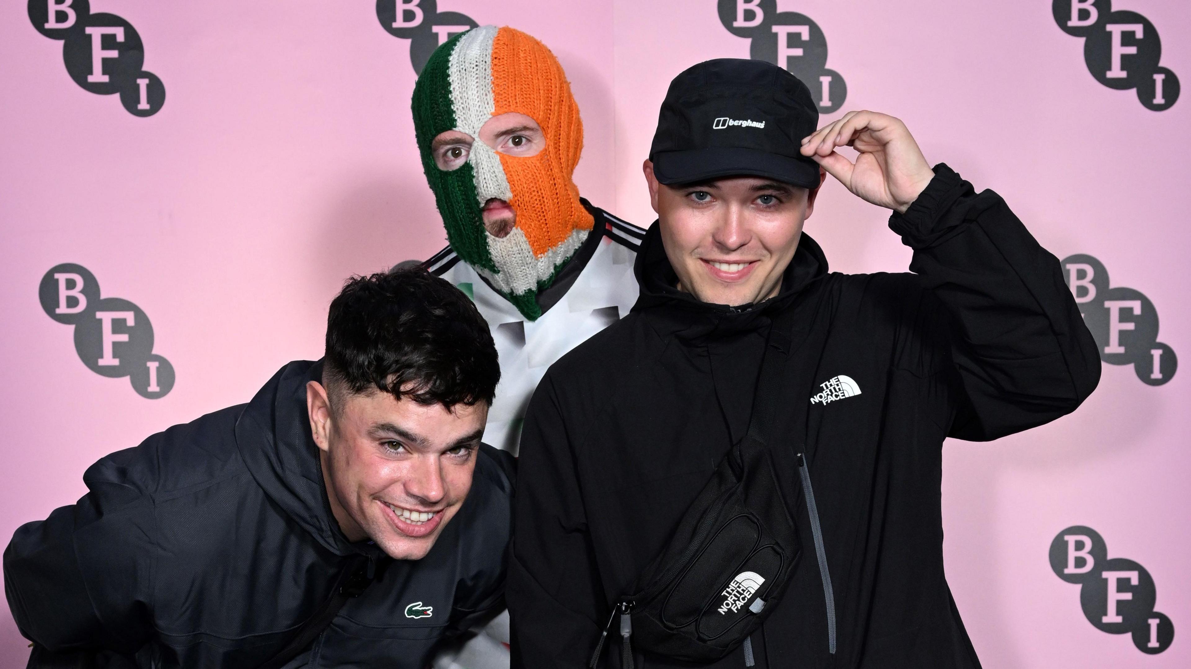 Three young men stand together. One is wearing a green, white and orange balaclava. A pink backdrop is behind them.