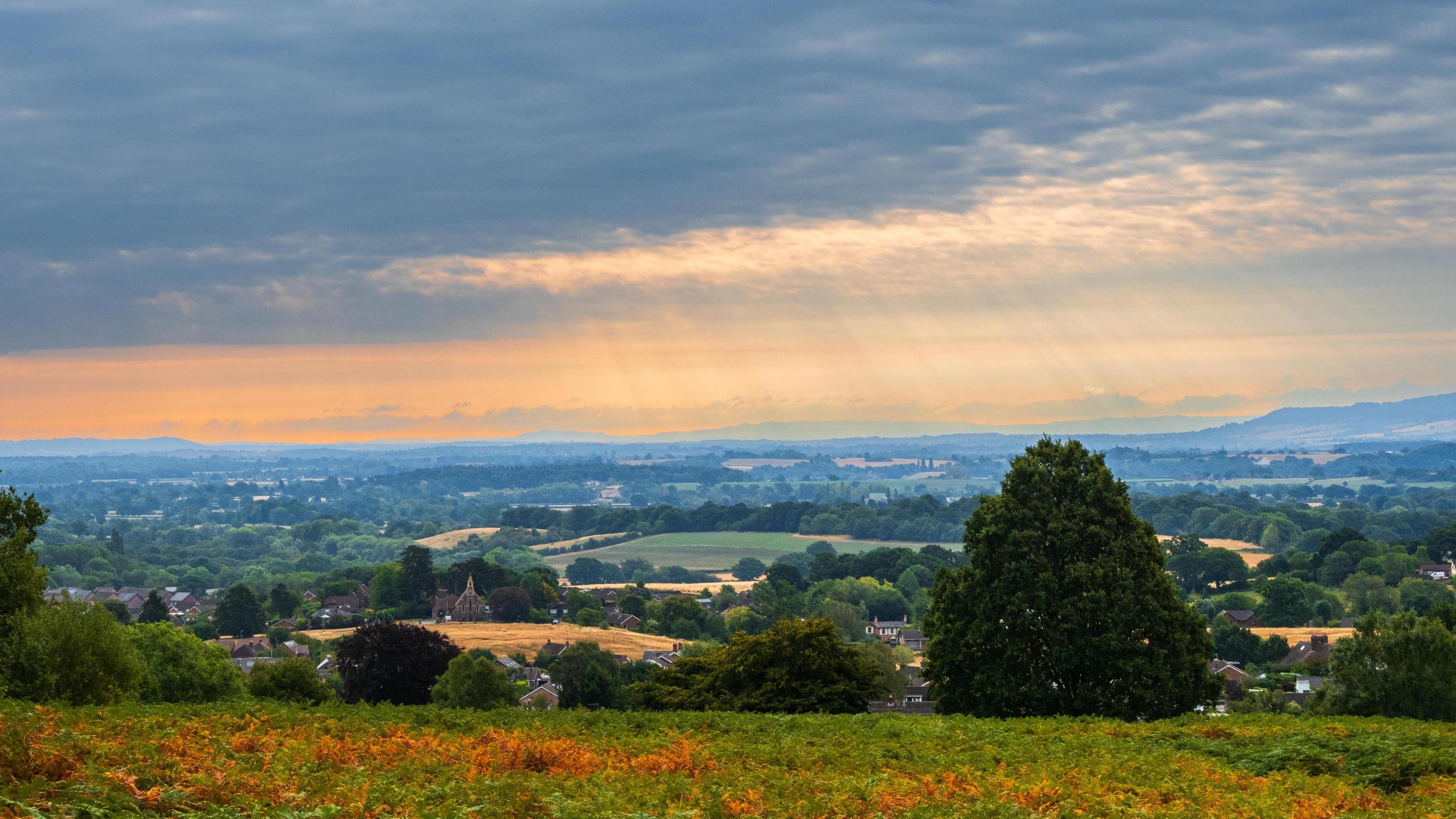 Sun rays through grey clouds above lush countryside with green fields and trees 