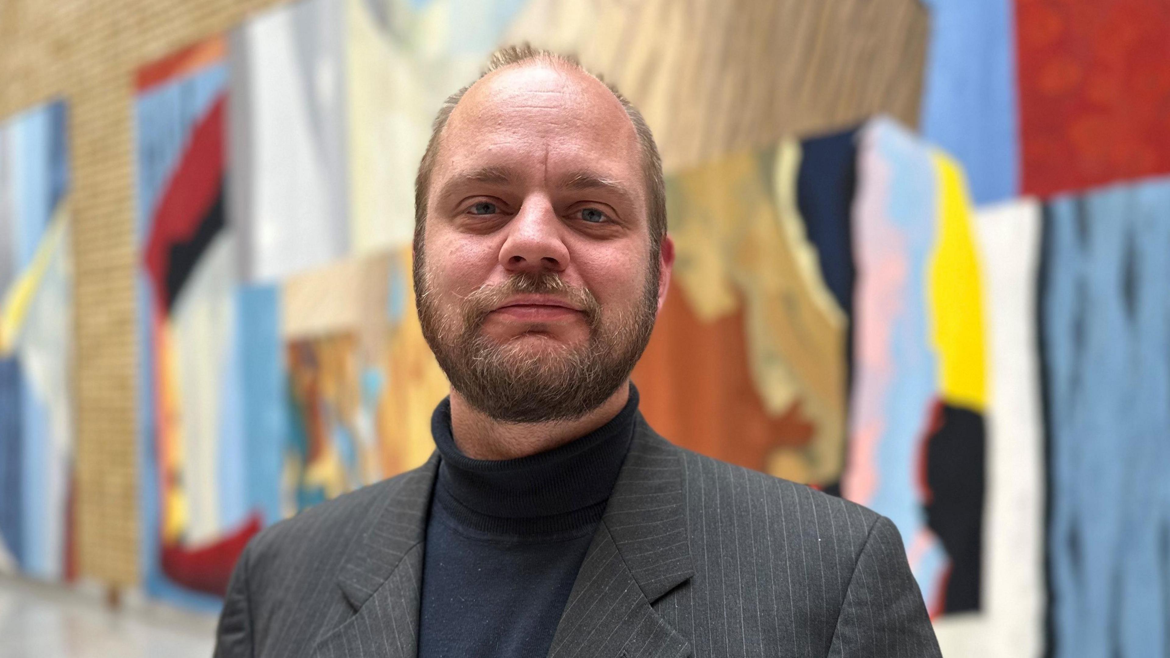 Mimir stands looking at the camera. He has short fair hair and a dark beard and is wearing a black turtleneck jumper with a grey suit jacket. Behind him is a large piece of colouful abstract art on a wall