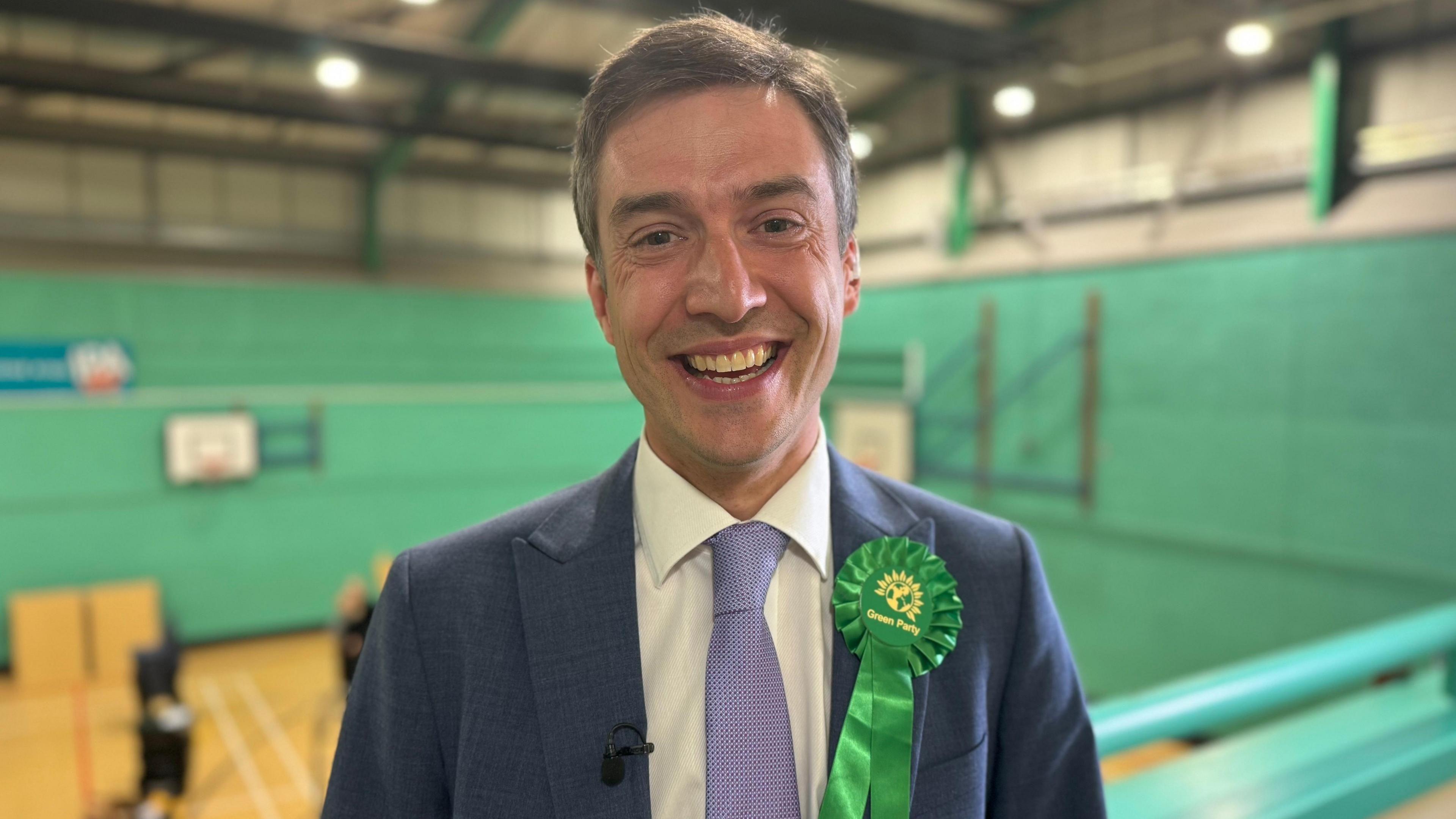 A smiling man with grey short hair wearing a white shirt, blue blazer and light blue tie. He also has a green Green Party rosette attached to his blazer. 