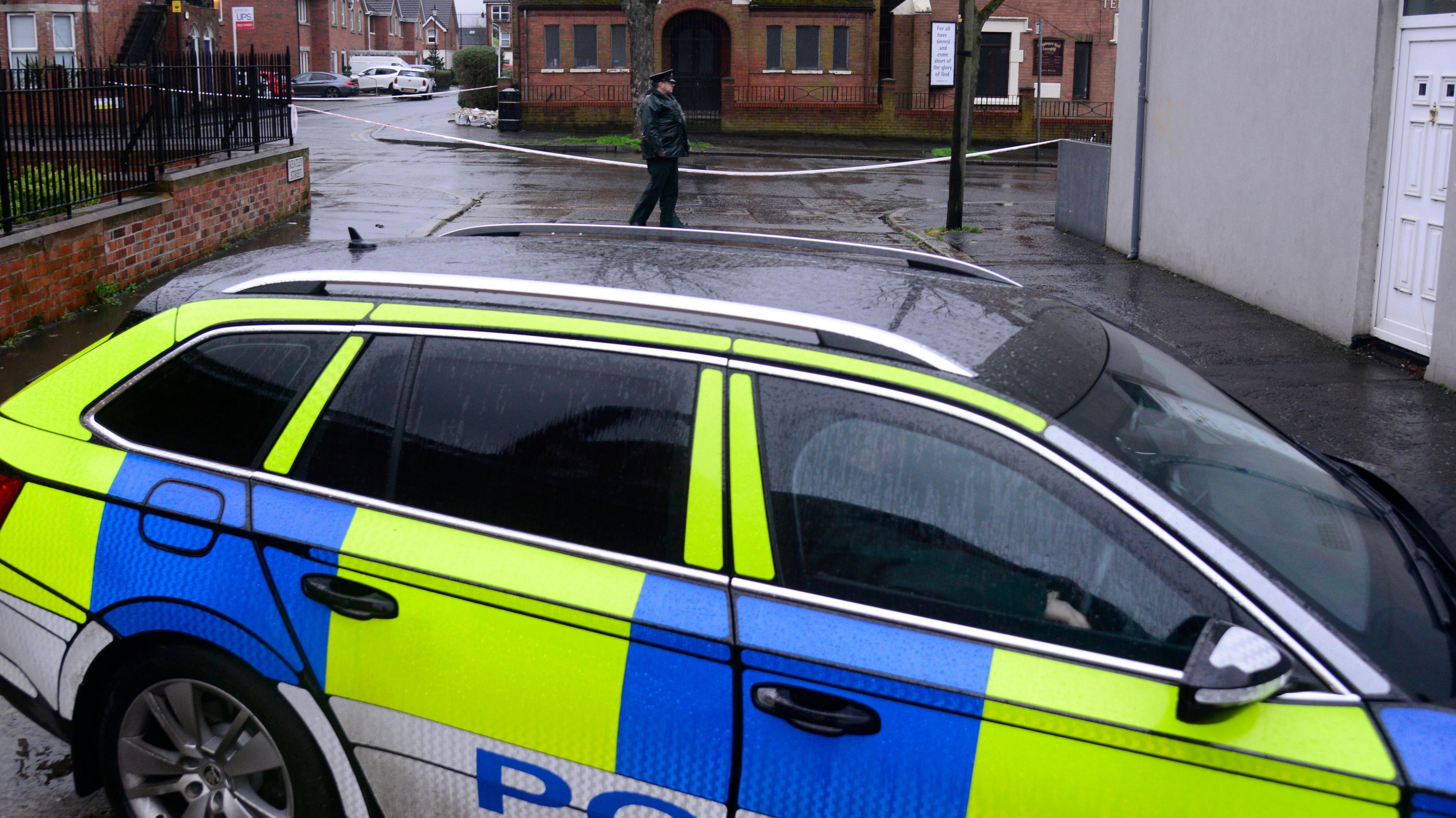 A yellow and blue police car parked beside police tape. A police officer can be seen standing in the background.