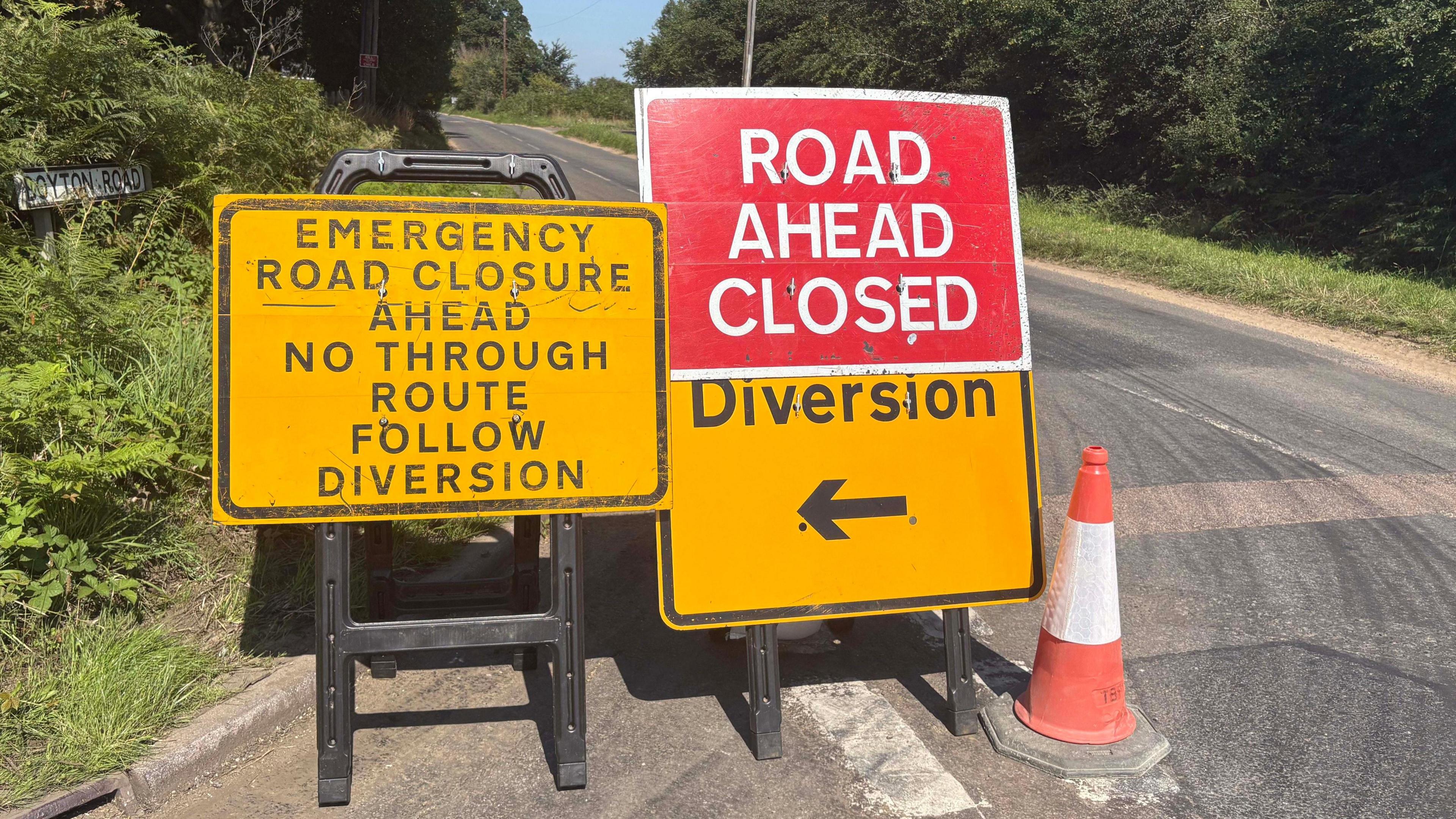 Sign reading: "Emergency road closure ahead no through route follow diversion". A second sign reads: "Road ahead closed". A third sign says "Diversion". The signs are on a road junction with bushes and trees either side of the road.