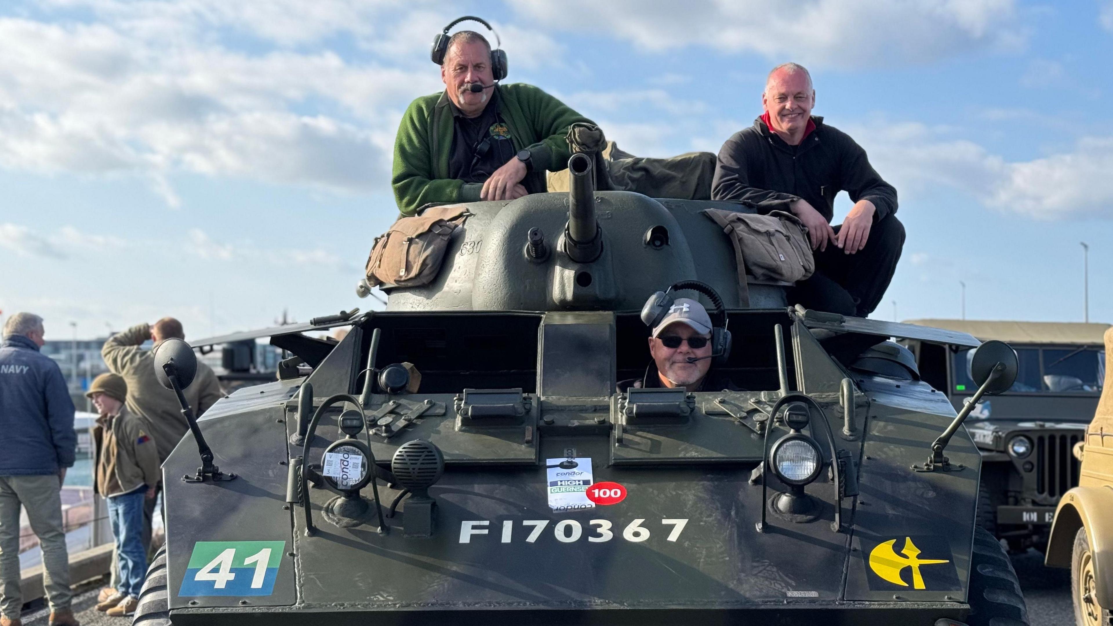 Three men sat on and in a small military tank. One man is peering out the window, while two men are sat on the turret.