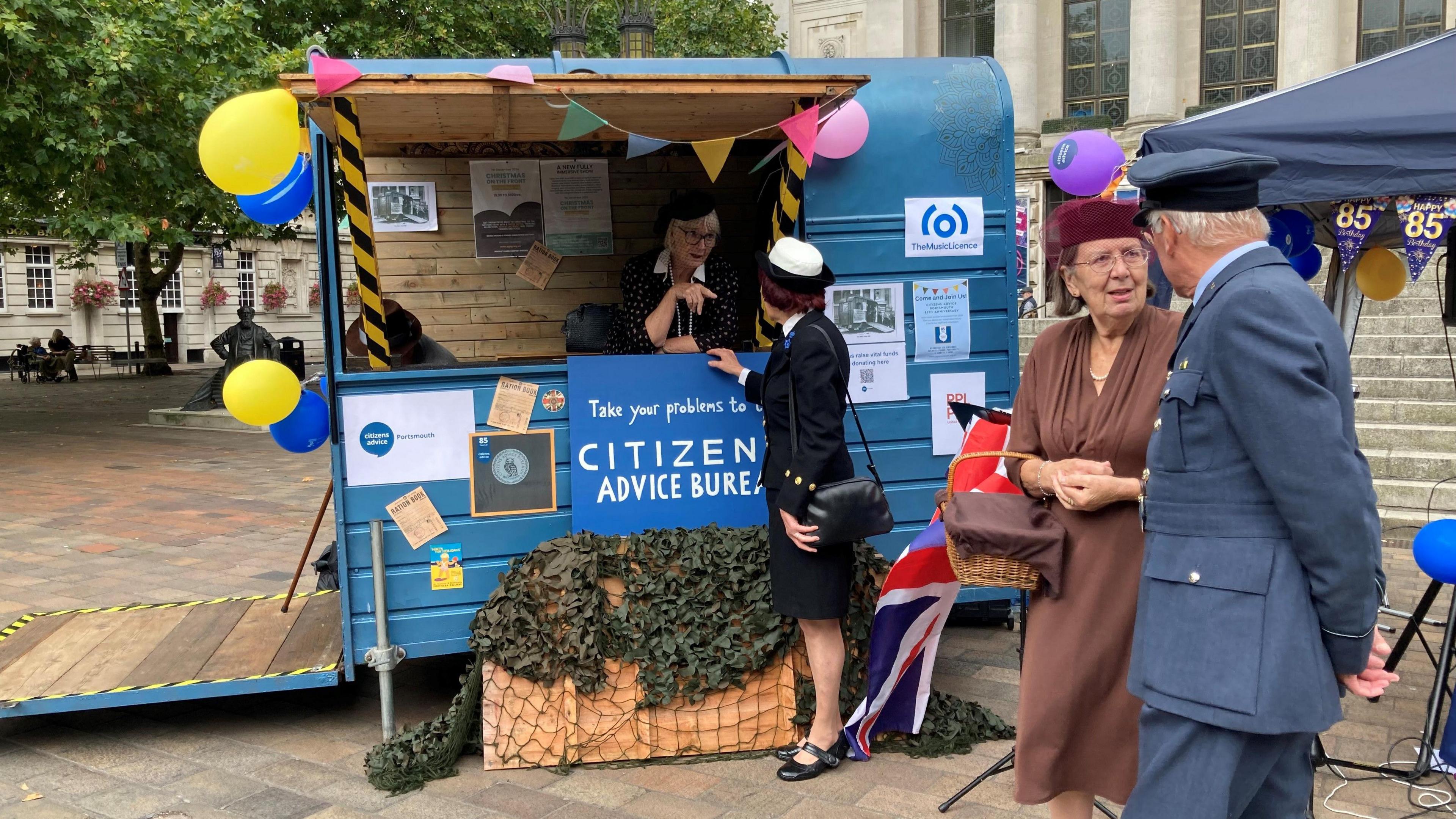 A modern photo of an old-fashioned looking horsebox with a serving counter, painted blue, with a sign saying "Citizens Advice Bureau". The woman in the horsebox, the woman she's talking to and the man and woman standing next to it are all dressed in period costumes. There are balloons and flags saying "happy 85th birthday" on the horsebox.