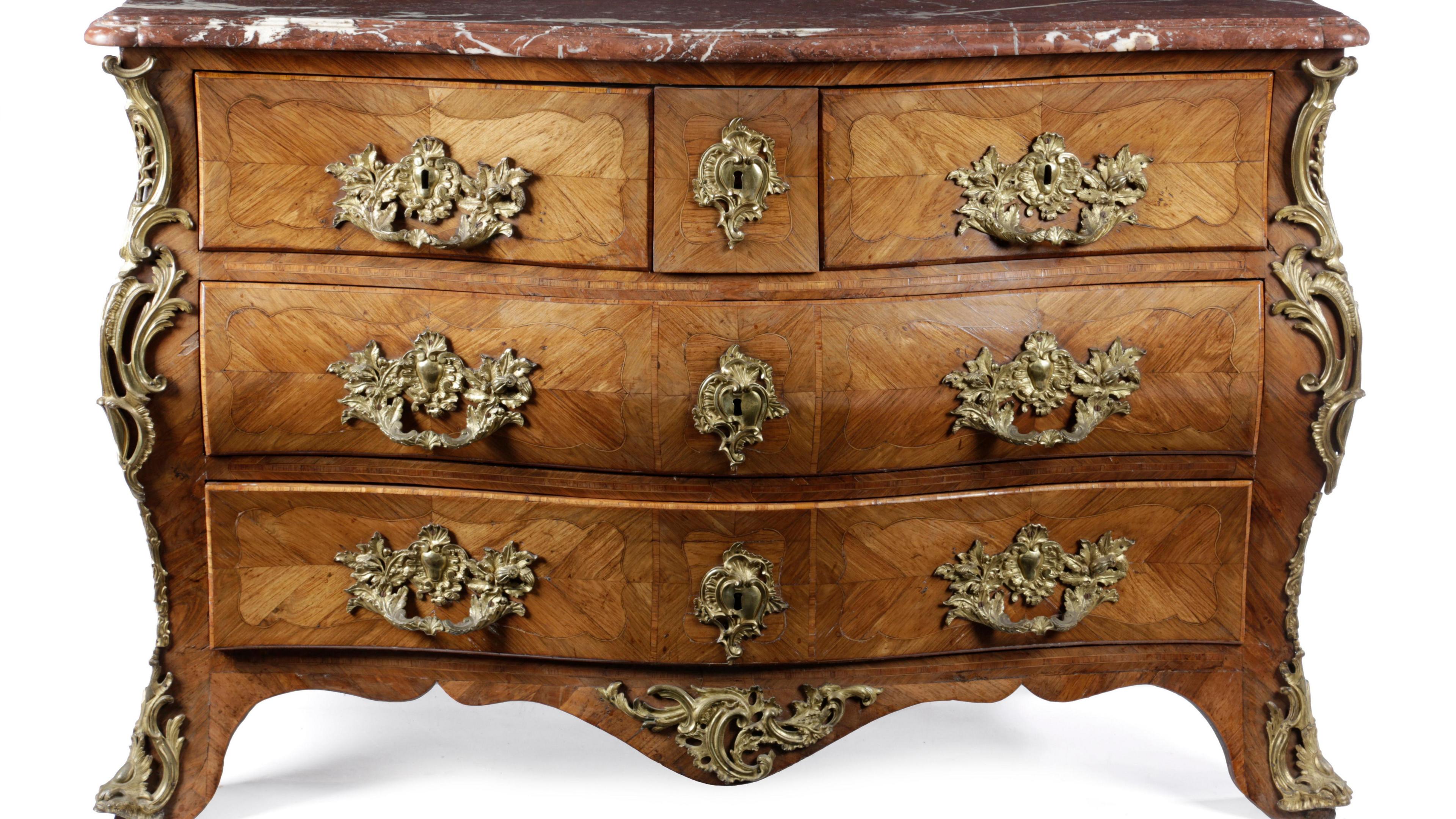A detailed and elaborate chest of drawers - a honey-coloured wood on feet carved with slight curves down the sides. It has a thick red marble top. There are two wide bottom drawers and three smaller at the top. Carved gold decoration is down the legs and sides. The handles are very elaborate and also in gold. 