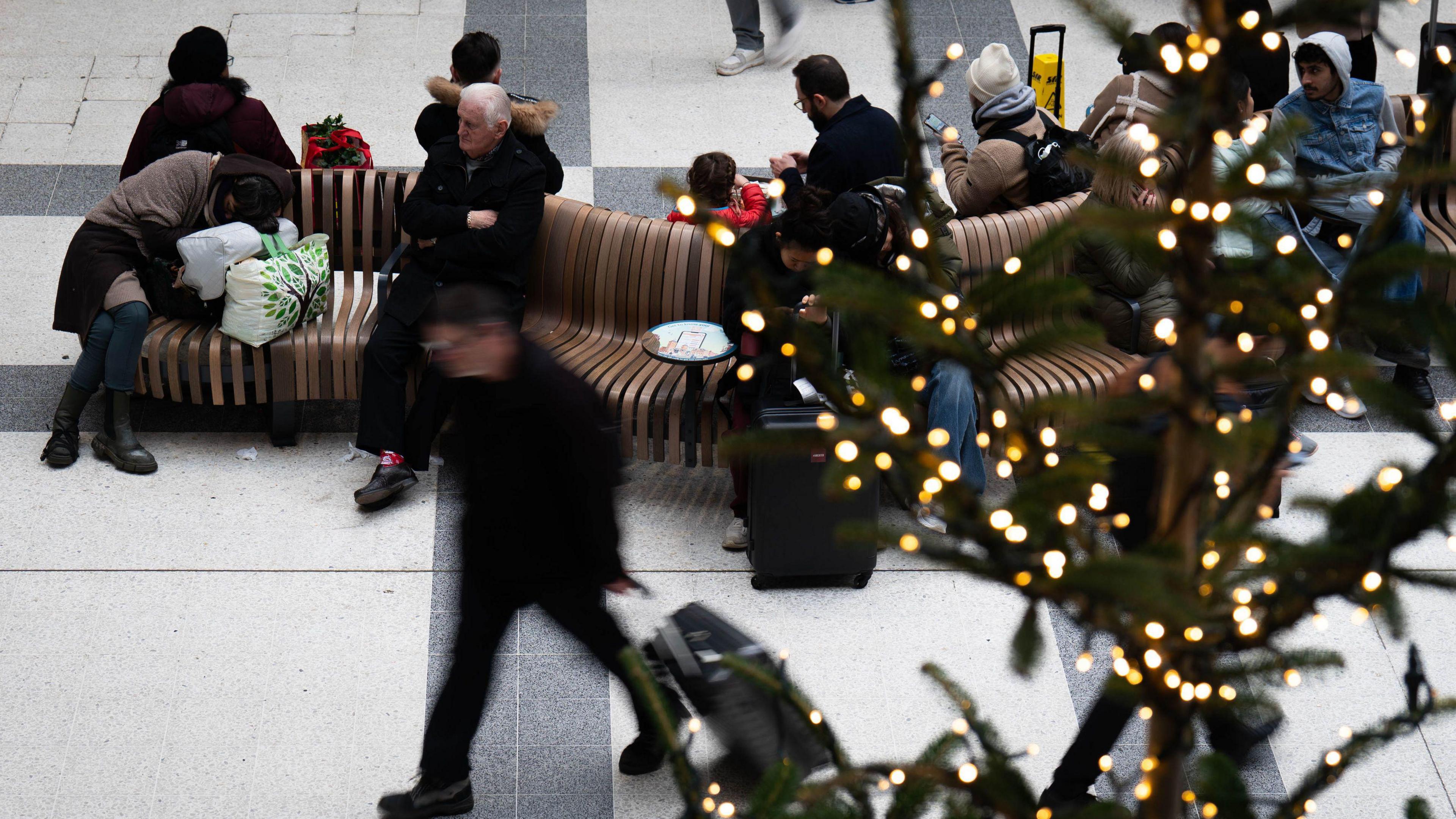 Station concourse. Some people are sitting on benches whilst others walk past with cases. There is a Christmas tree with white lights in the foreground.