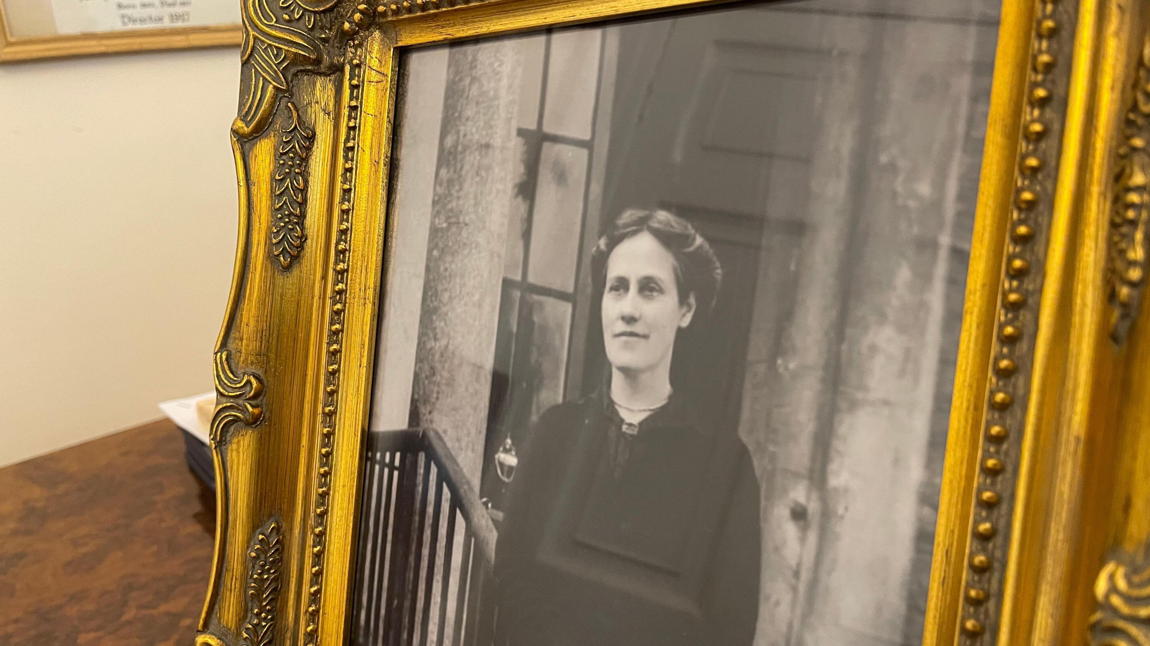 A framed black and white photo of Theresa Hardcastle. She has long hair which has been parted and tied up around her head. She is wearing a black dress and is standing on the steps of a large building, leaning one arm on a bannister.