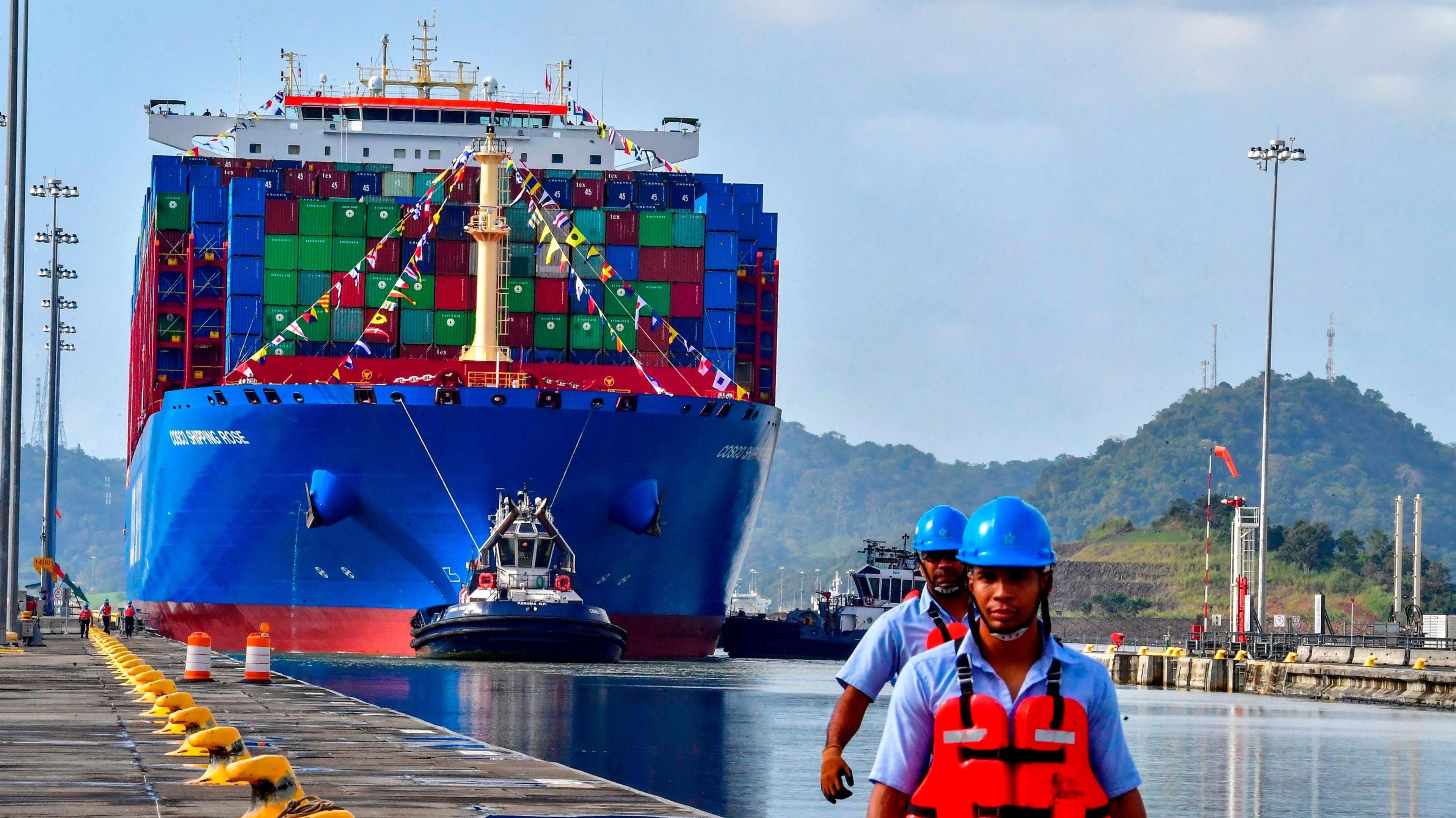 A bright blue container ship sails along the Panama Canal. Two workers in blue helmets and orange hi-vis jackets stand in the foreground.