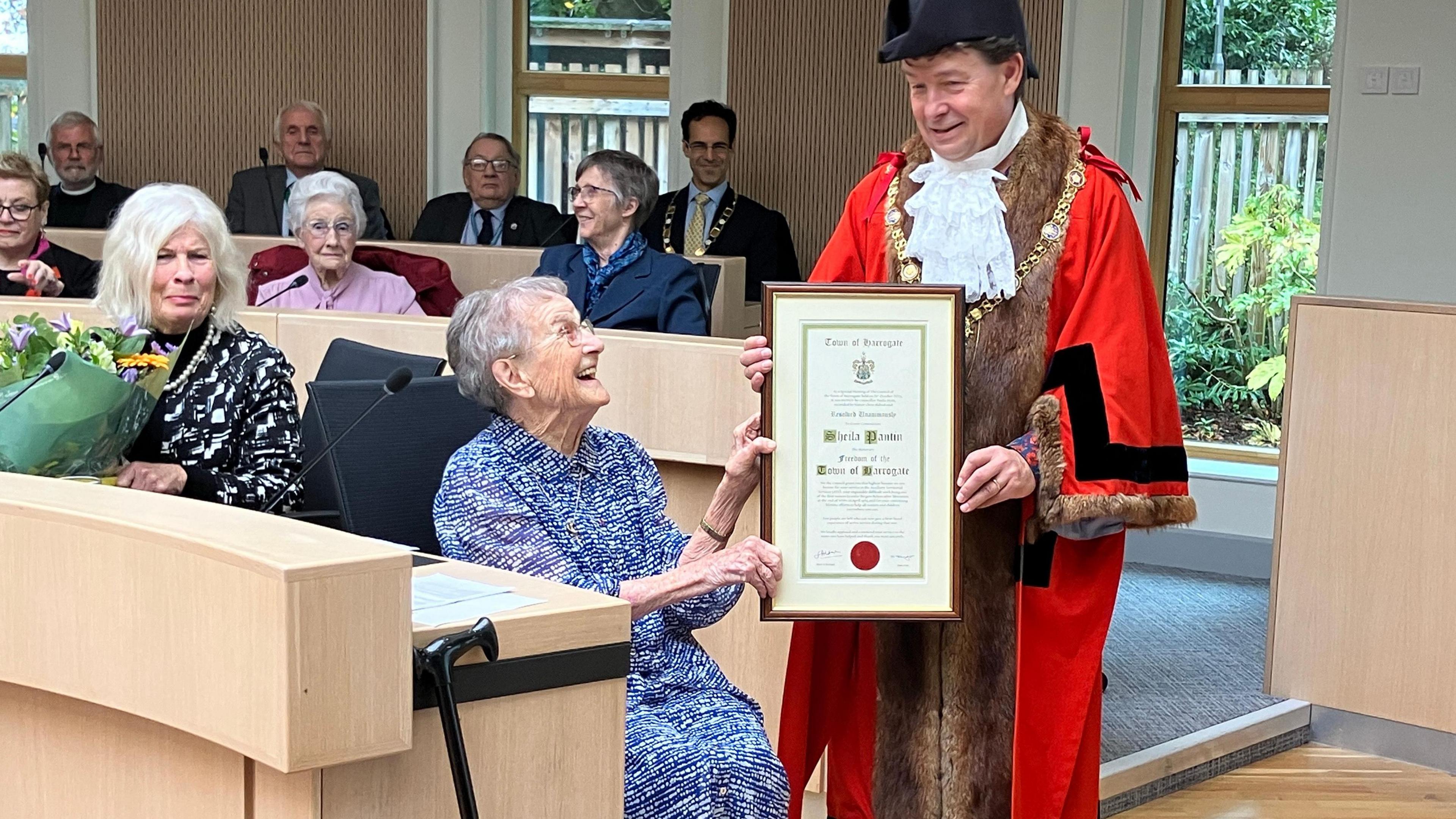 A meeting of Harrogate Town Council with the mayor in full ceremonial dress holding a framed scroll awarding the freedom of the town to Mrs Sheila Pantin who is seated looking at the award in the mayor's hand