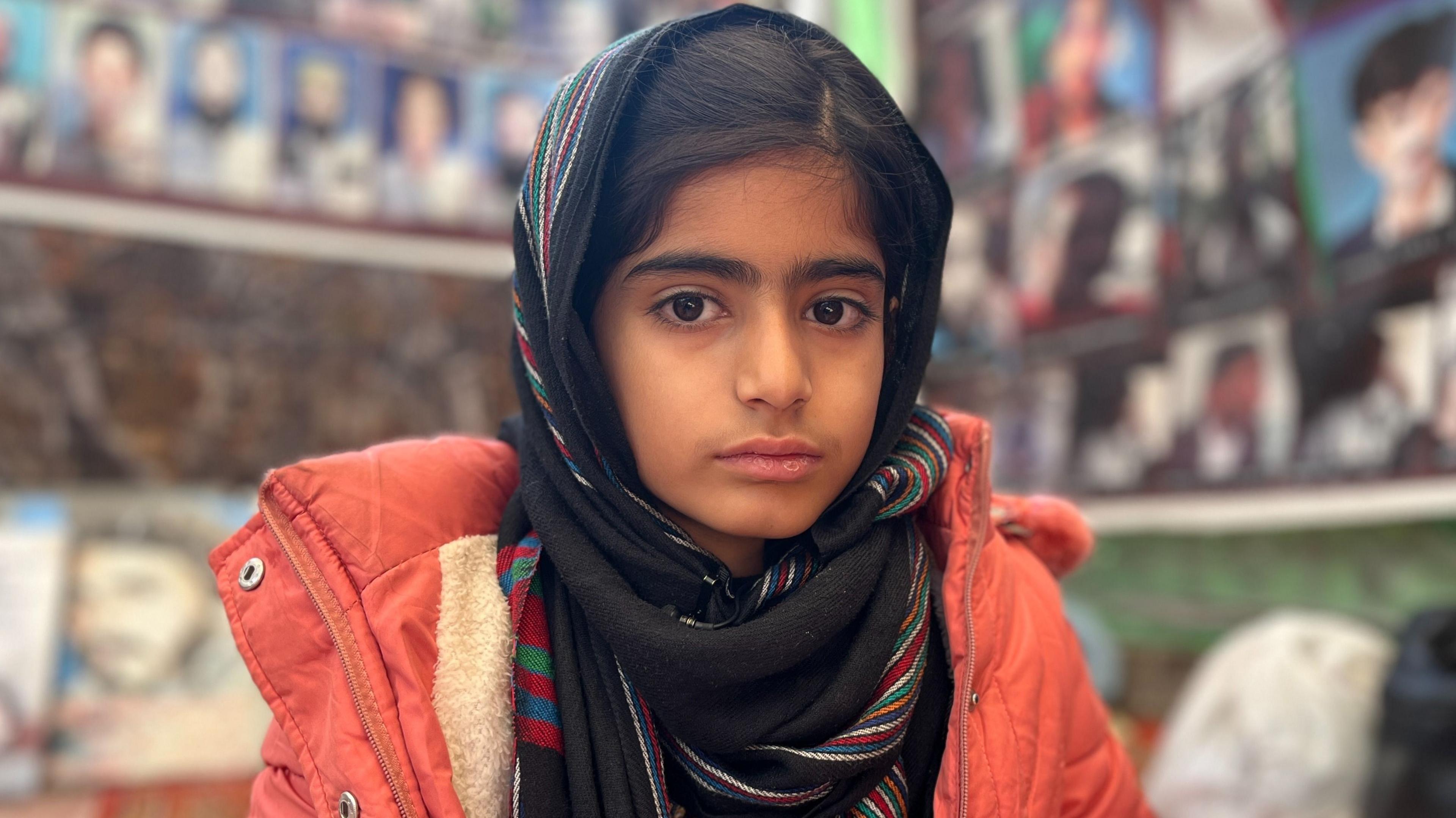 A young girl in headscarf and orange coat looks at the camera