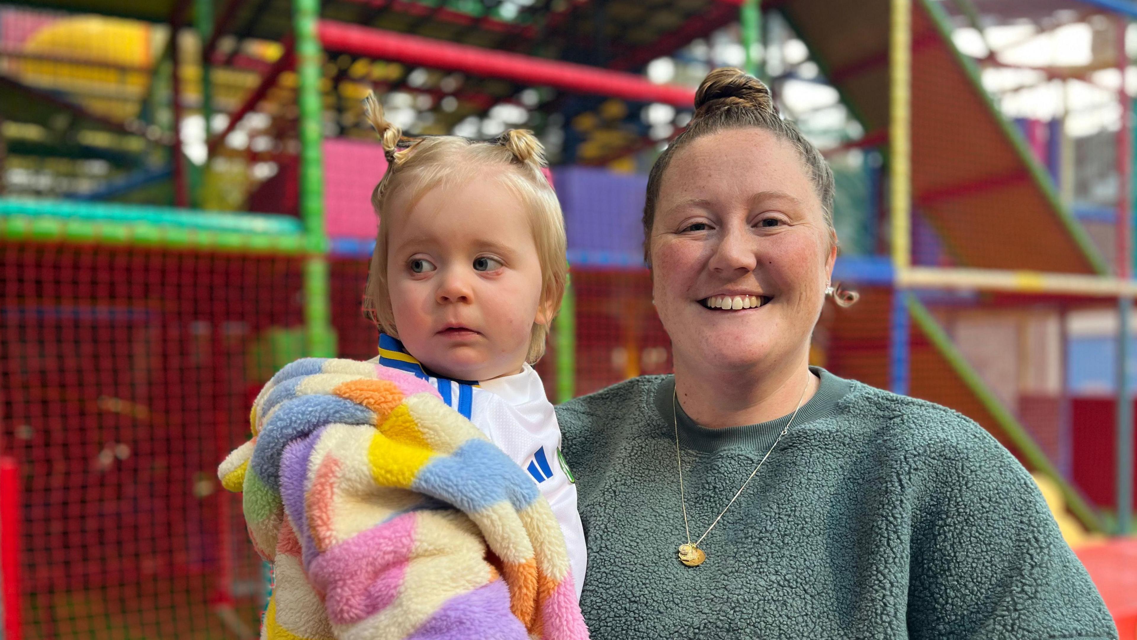 Lauren Byrne is smiling at the camera while holding her child Isla. They are in front of a colourful soft play area.