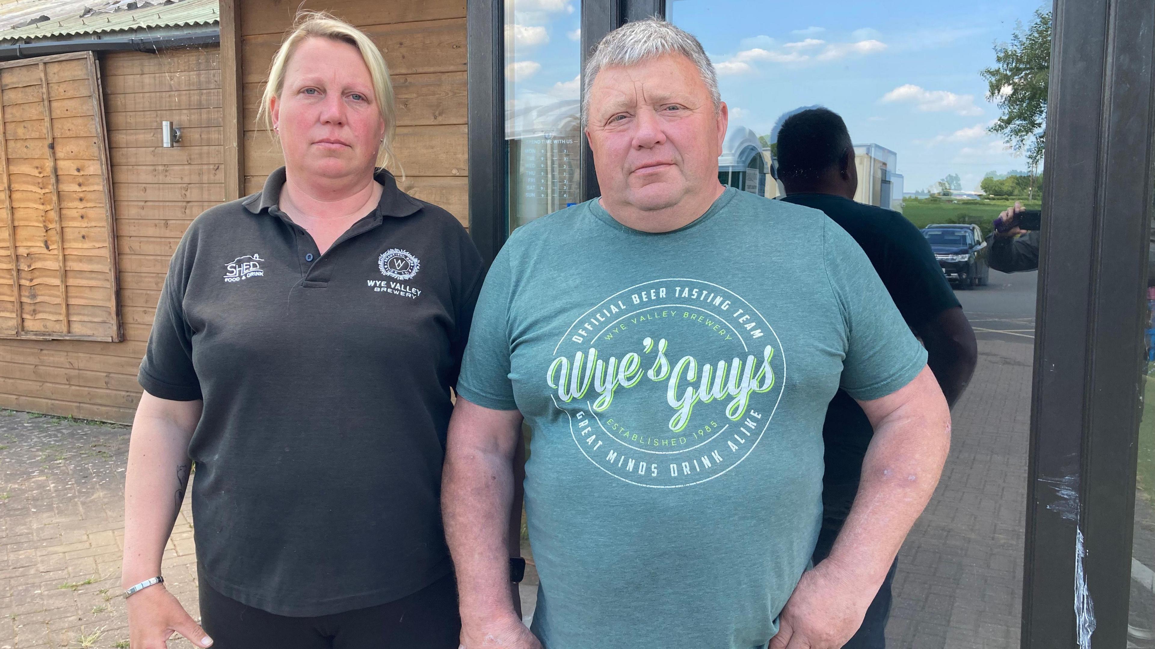 A woman with tied back blonde hair is wearing a black T-shirt and is stood in front of a wooden building. Next to her stands a man with grey hair wearing a green T-shirt with a large logo on.
