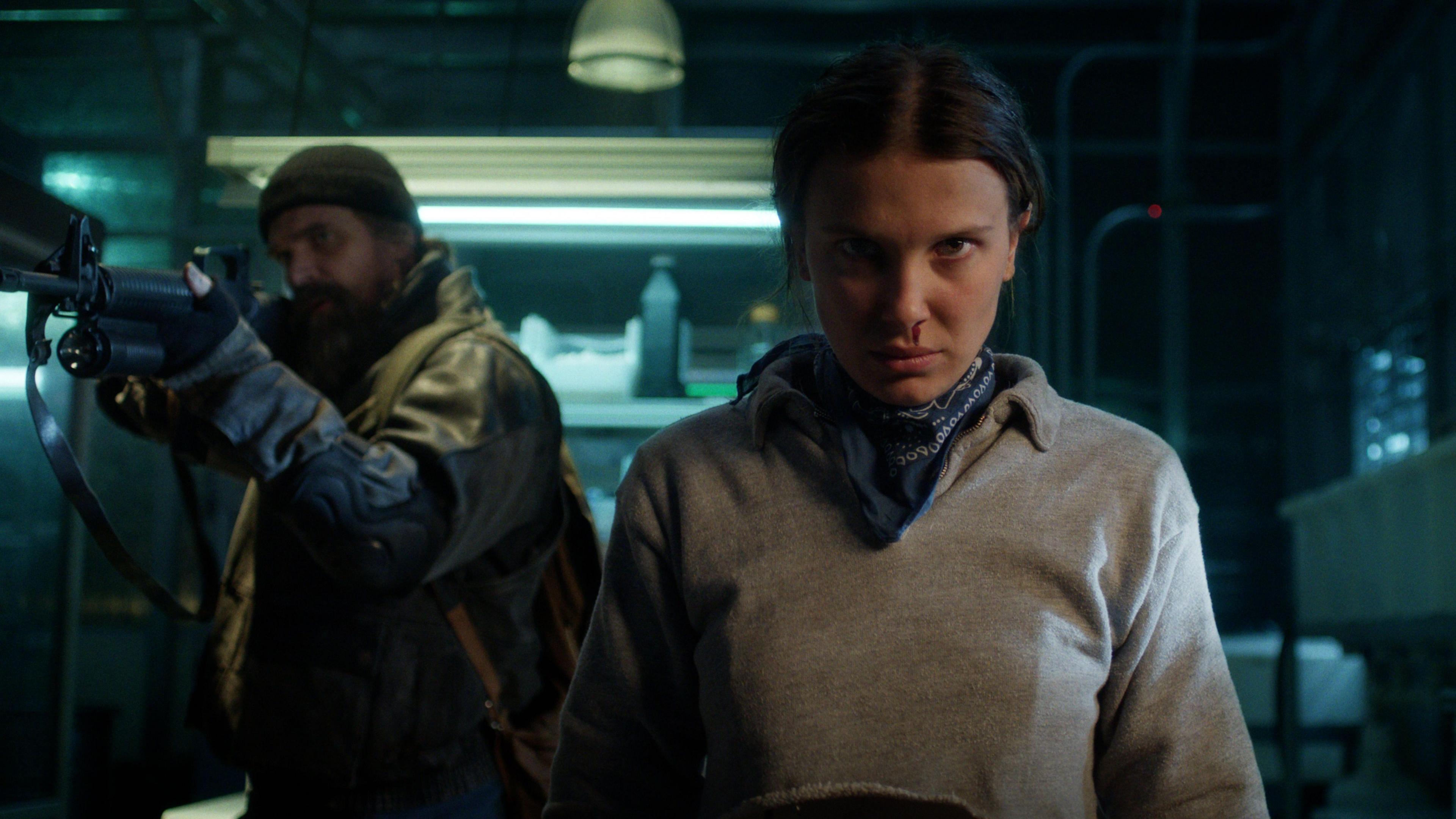 A still from a scene in season five of Stranger Things, with David Harbour poiting a rifle out of the frame, next to Millie Bobby Brown who stands with a focused expression and blood dribbling from one nostril