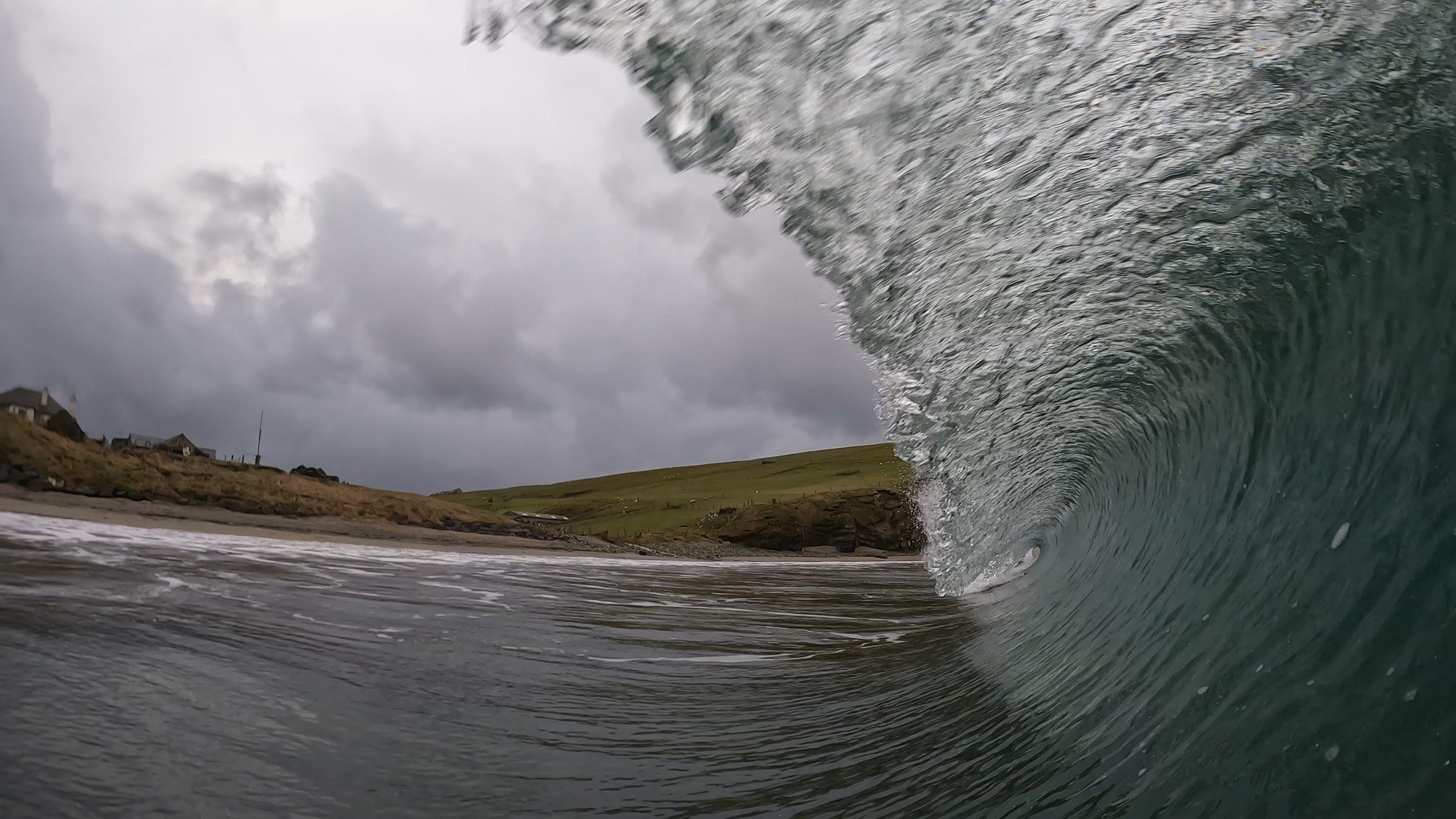 A powerful ocean wave curls dramatically, forming a tunnel of foamy water near the shore. The coastline in the background features grassy hills and a few scattered buildings under a stormy, grey sky.