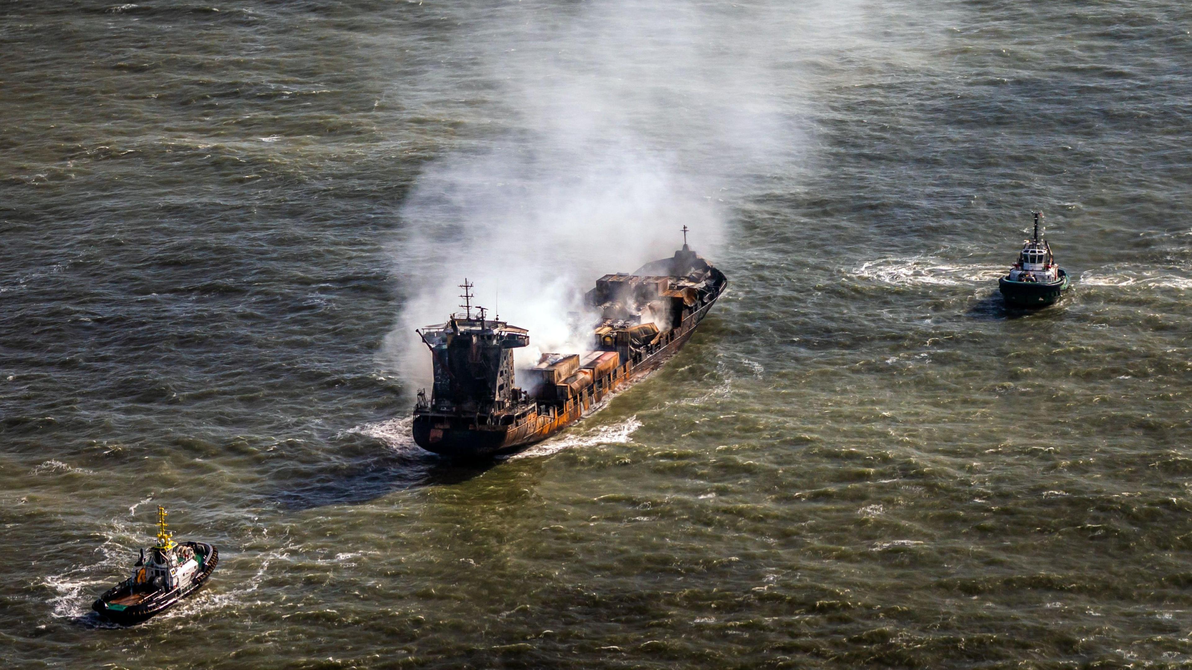 Grey smoke billowing from a cargo vessel adrift at sea with two tug boats either side of it – one in front and the other behind.