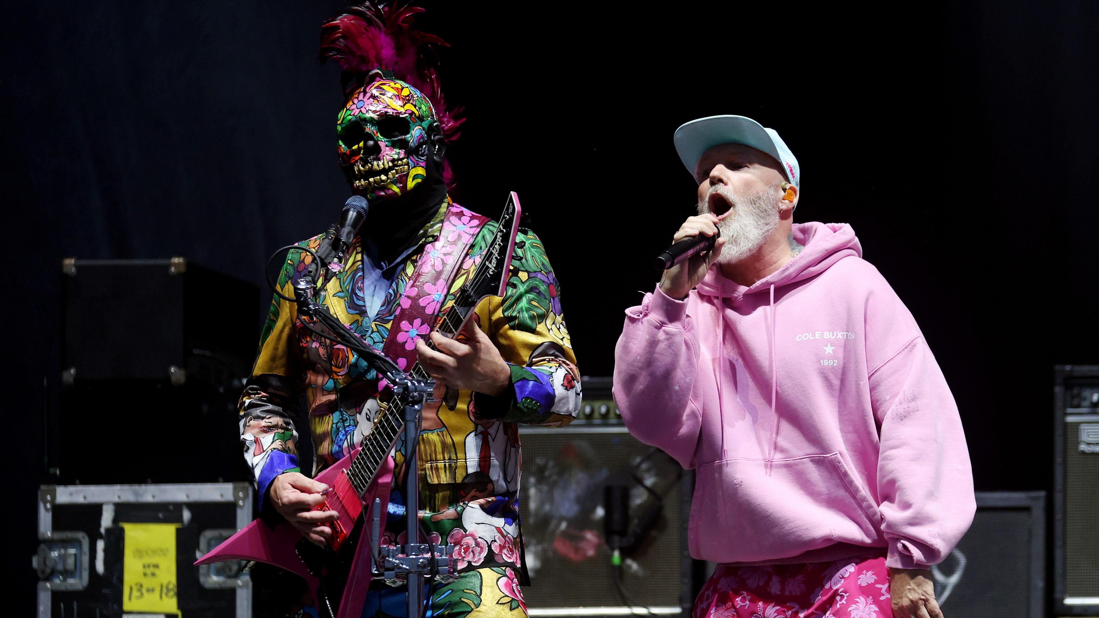 Wes Borland in a multi-coloured floral mask and suit playing a pink guitar and Fred Durst of Limp Bizkit singing wearing a pink hoodie and pink floral shorts, with white cap on his head