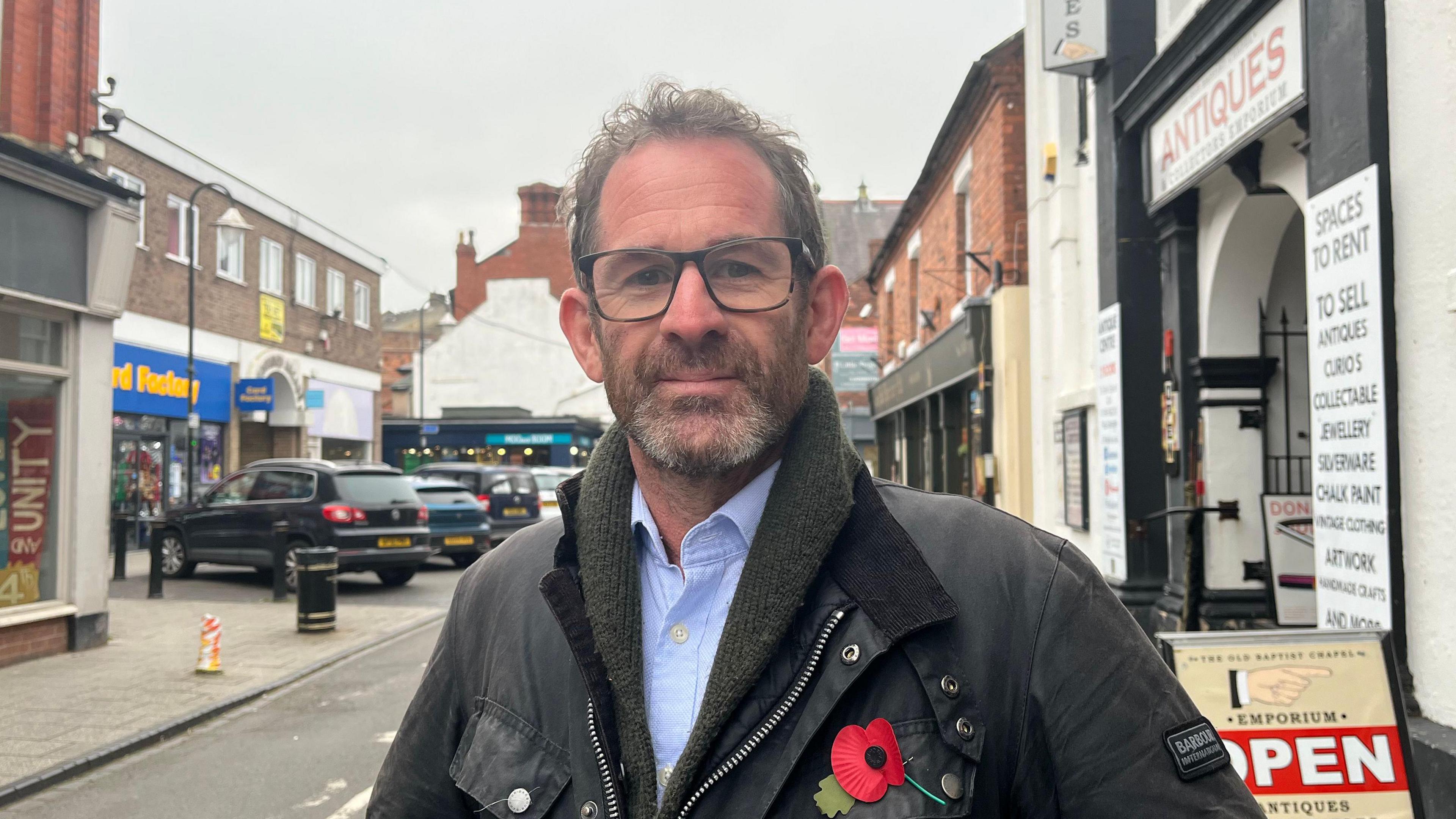 A man with curly dark hair is smiling slightly and looking into the camera. He's wearing a blue collared shirt, a dark green scarf and a dark brown wax jacket. There is a red paper poppy pinned to his lapel. He's stood on a street with shops just out of focus in the background. The sky is grey and overcast.
