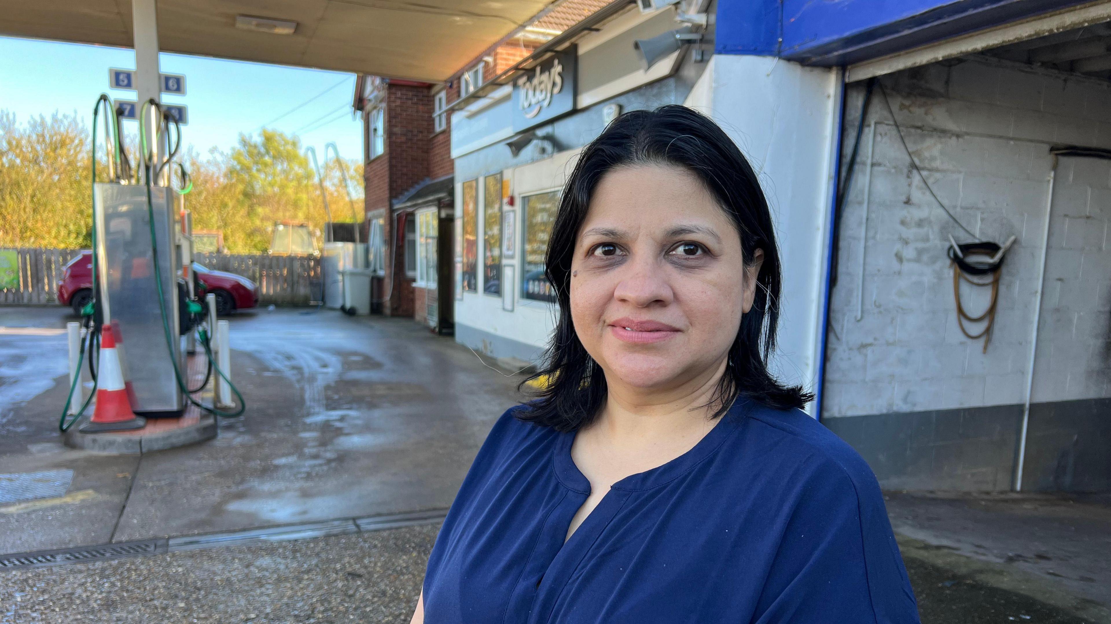 A head and shoulders shot of a woman with black hair, wearing a dark blue blouse. She stands on the forecourt of a petrol station, with pumps, a shop and a garage area in the background.