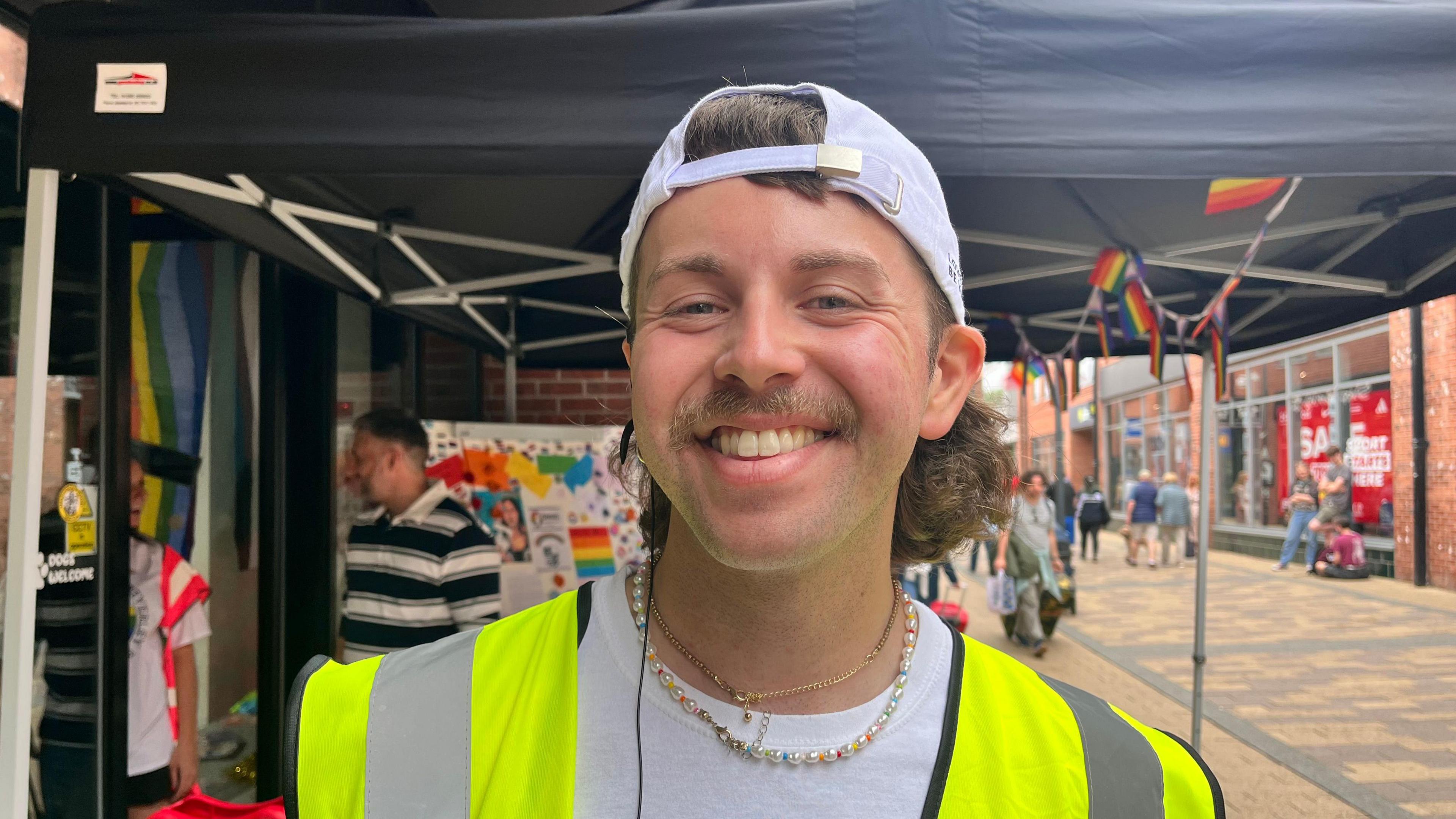 A head and shoulders shot of Dan Rothery. He has a light brown moustache and a light brown mullet. He is wearing a white cap backwards on his head, a pear necklace, a white t-shirt and a yellow hi-visibility vest. He is stood in front of a black canopy on what looks to be a High Street.