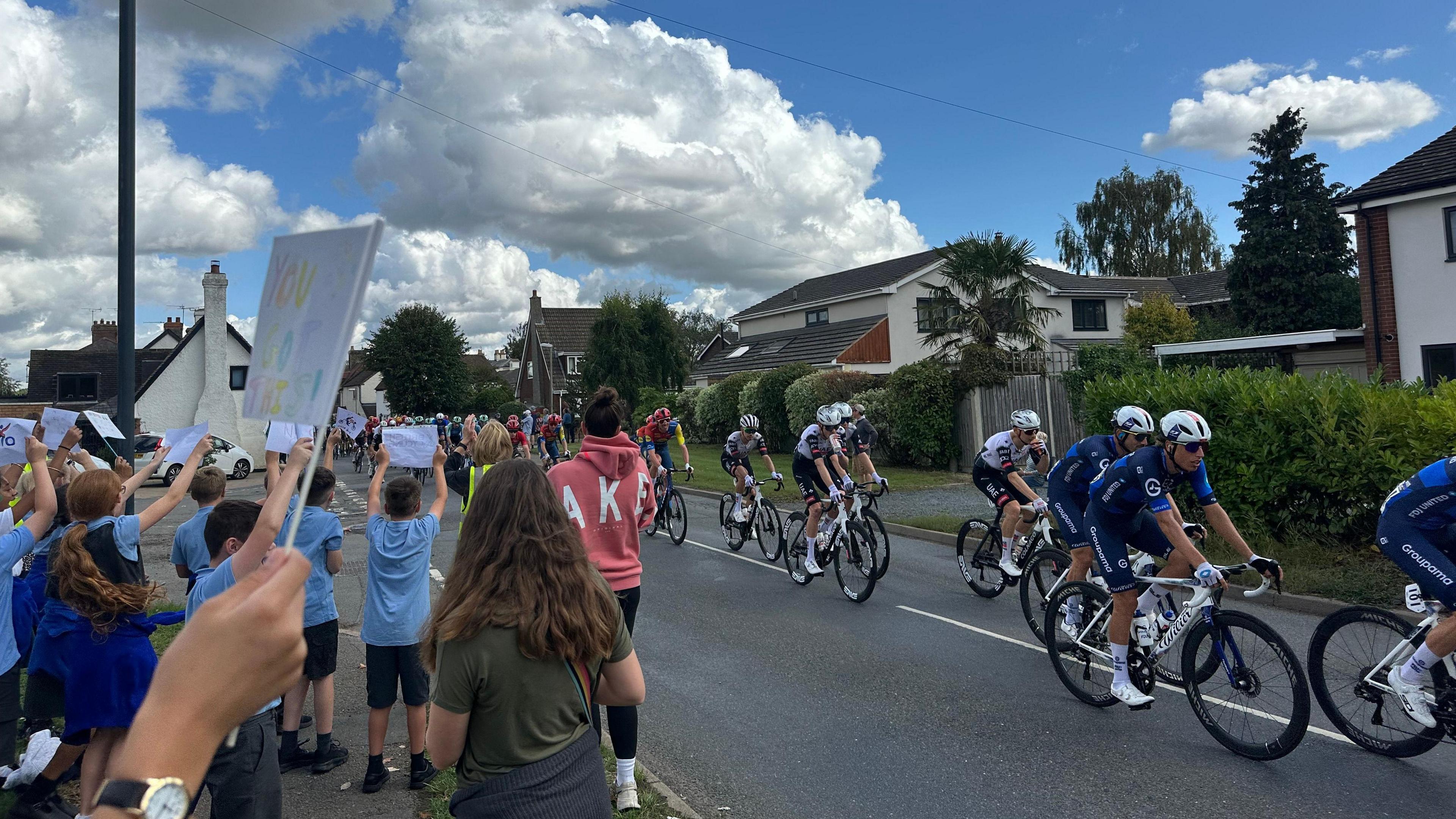 Schoolchildren and the public line the streets of Warwickshire as riders cycle past. Fans are holding up banners. The race is passing through a rural area with houses and gardens in the background.
