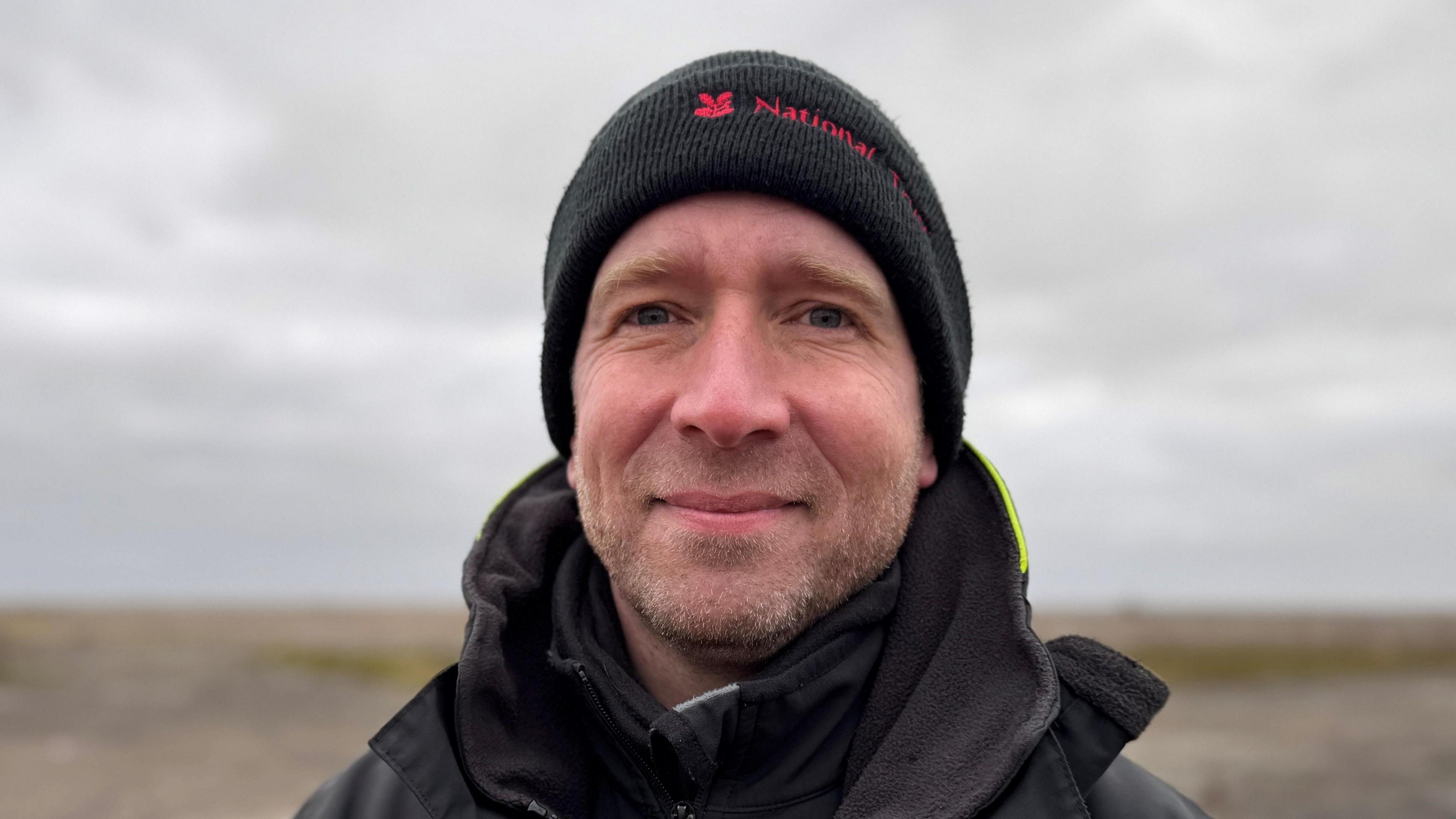 Matt Wilson smiles at the camera while standing on a shingle beach. He is wearing a black coloured beanie hat and a black coat. He has light coloured stubble.