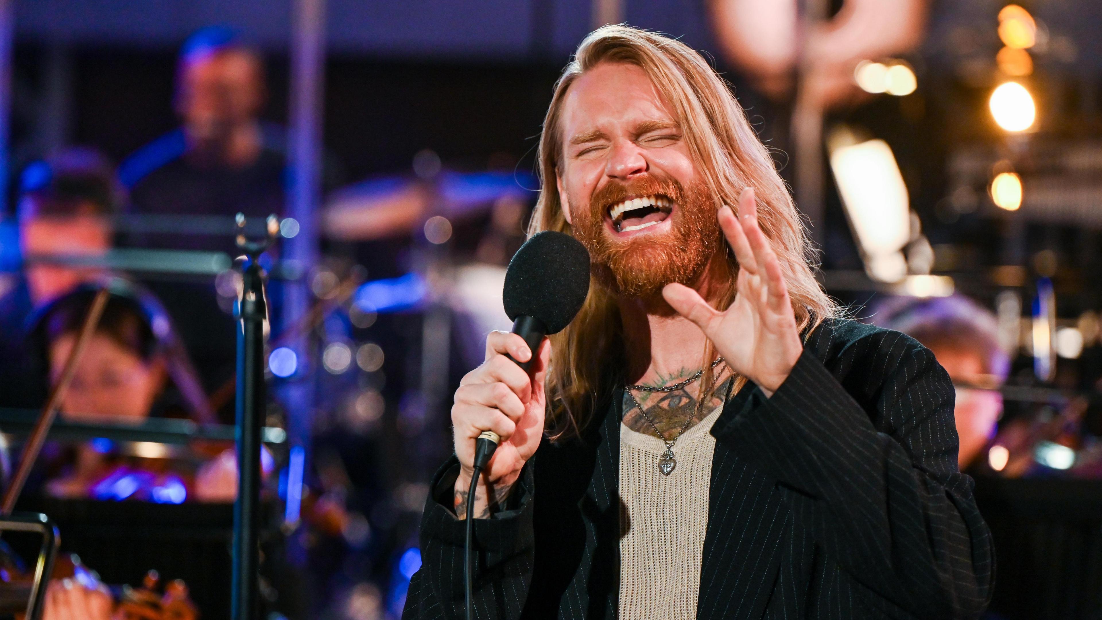 Sam Ryder sings into a microphone, with members of an orchestra out of focus behind him. He has long flowing blond hair and is wearing a black pinstripe jacket.