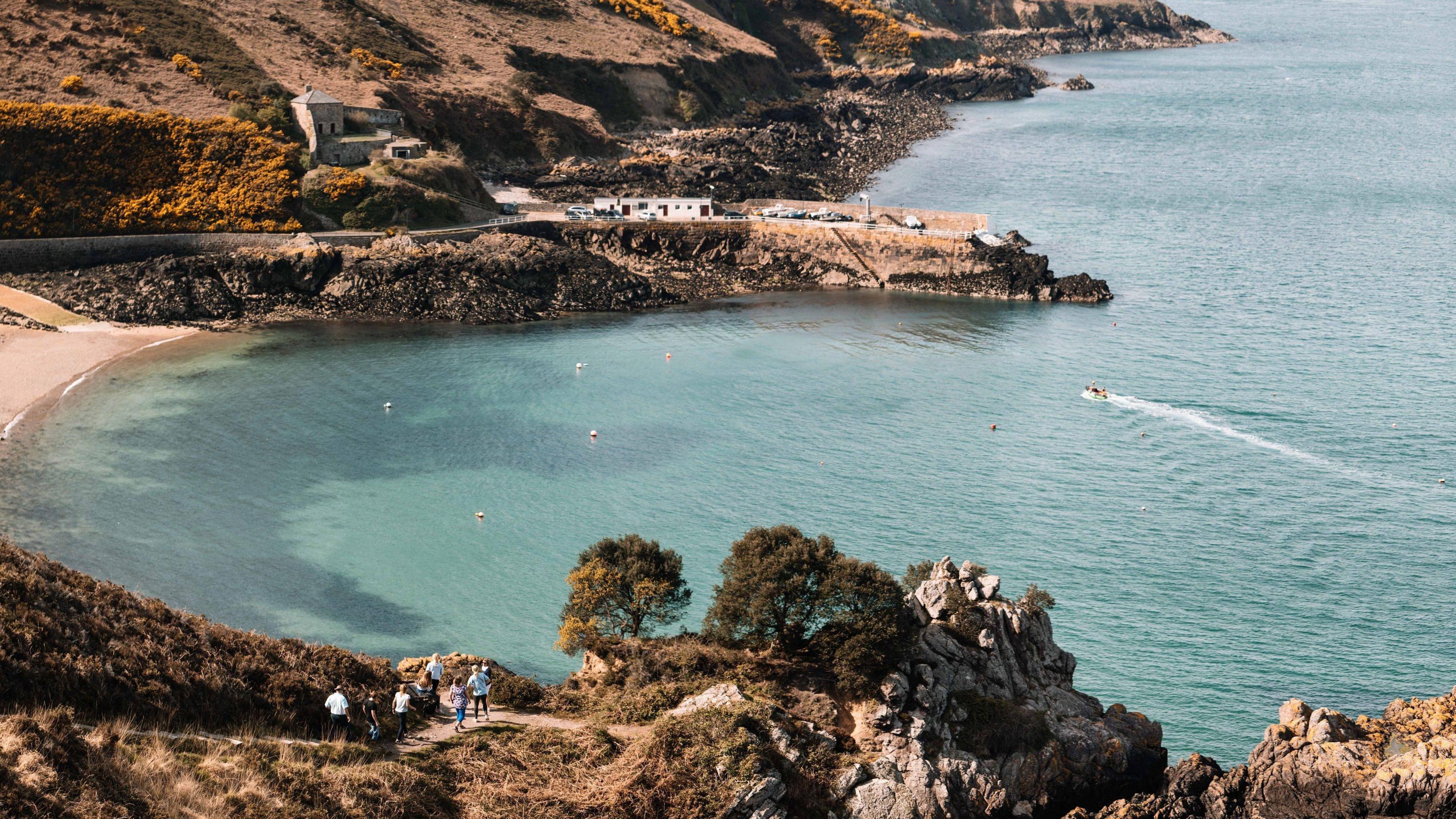 Walkers on cliffs above a bay in which there are swimmers and a boat