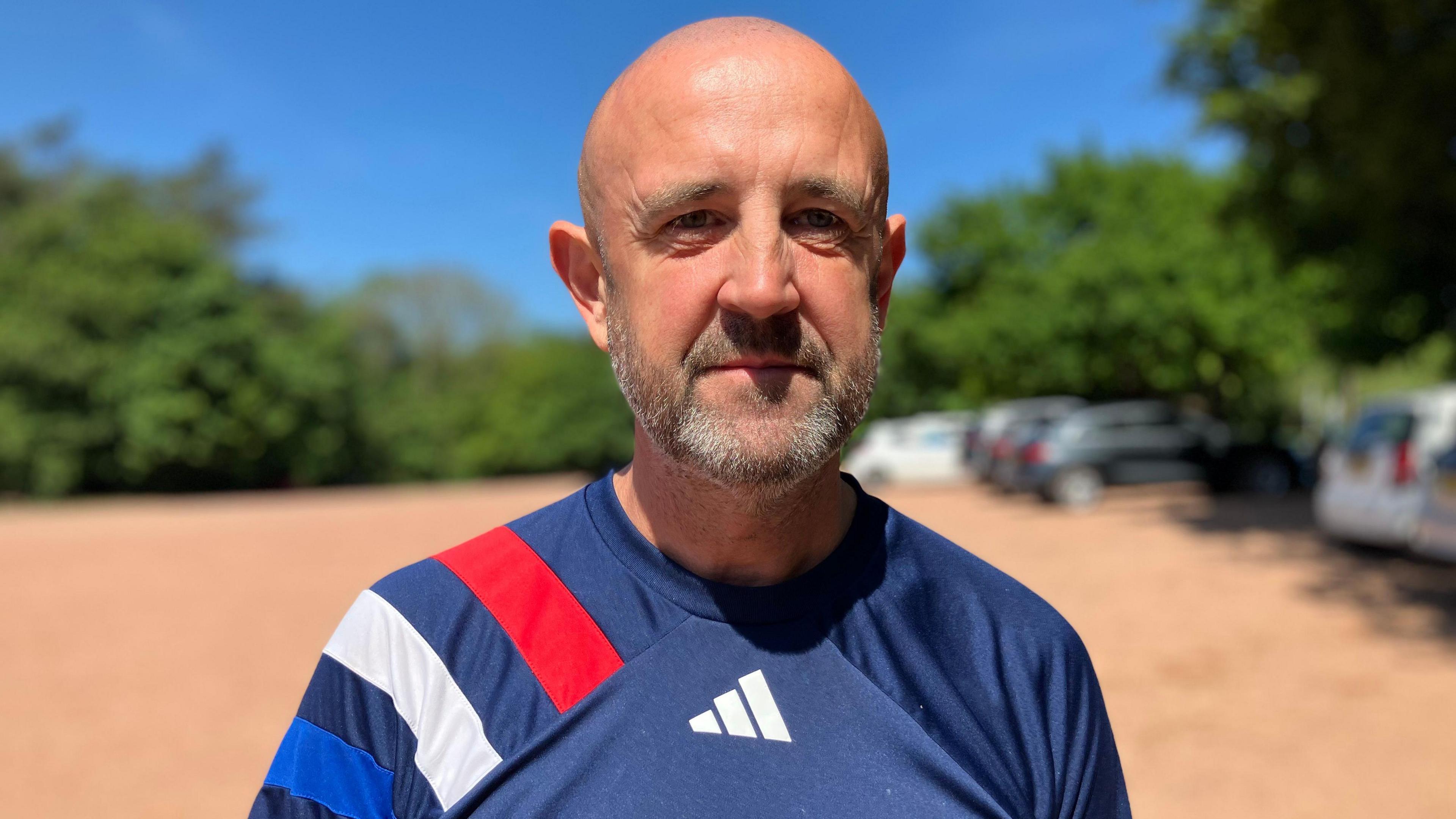 A man stood in a large car park. He is bald, has a grey and black beard and is wearing a blue Adidas sports top with red blue and white stripes on his shoulder. There are cars parked on a red gravel car park behind him.