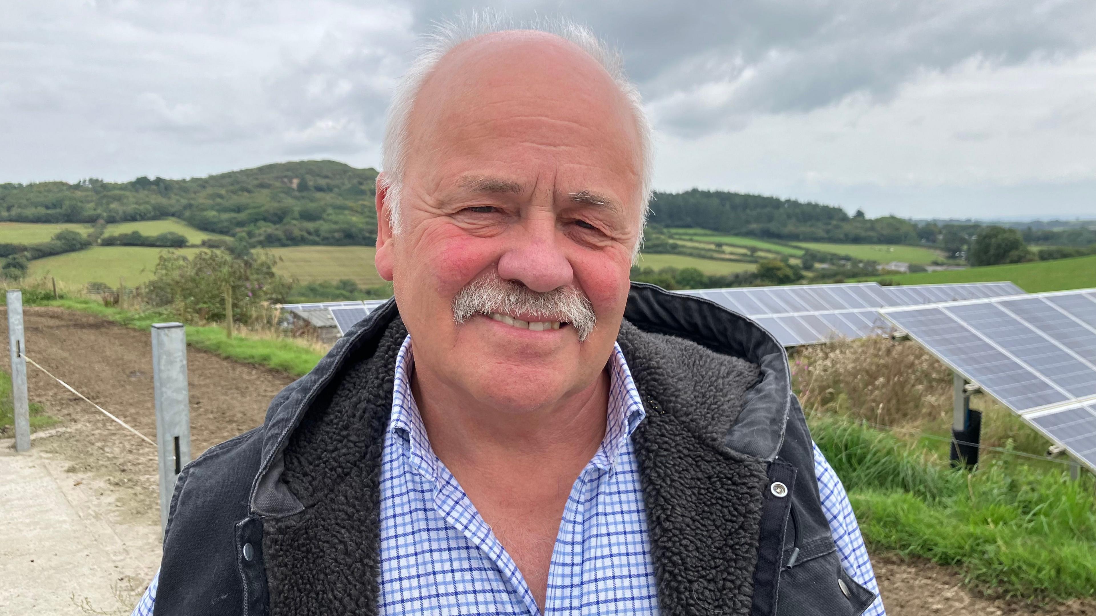 Charles Baughan, a Taste of the West director, standing in front of fields and solar panels. He is wearing a blue checked shirt and a dark grey hooded gilet. He has a white moustache and is bald