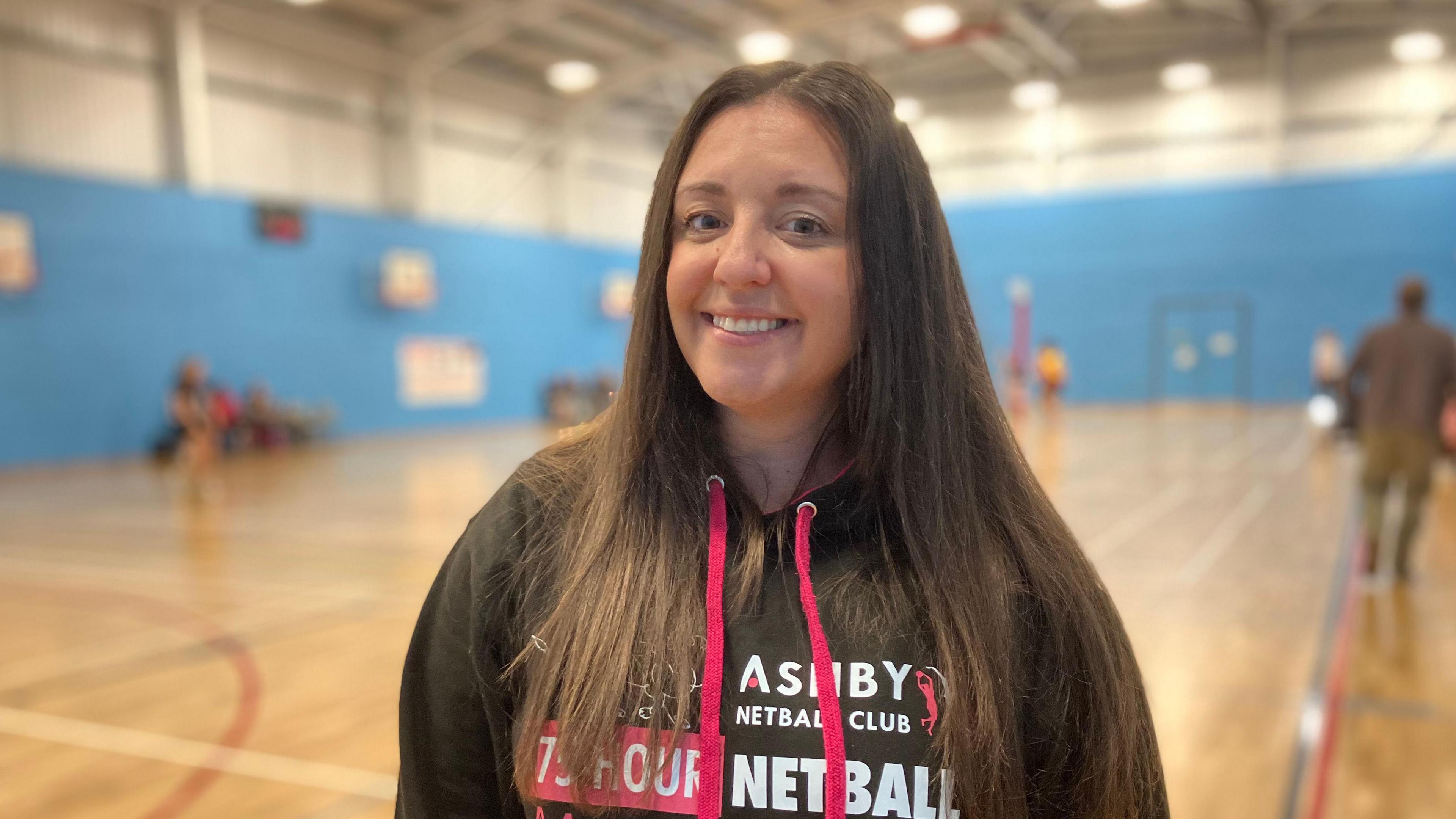 A woman with long dark hair, wearing a black hooded top smiles at the camera.  She stands in front of a netball court