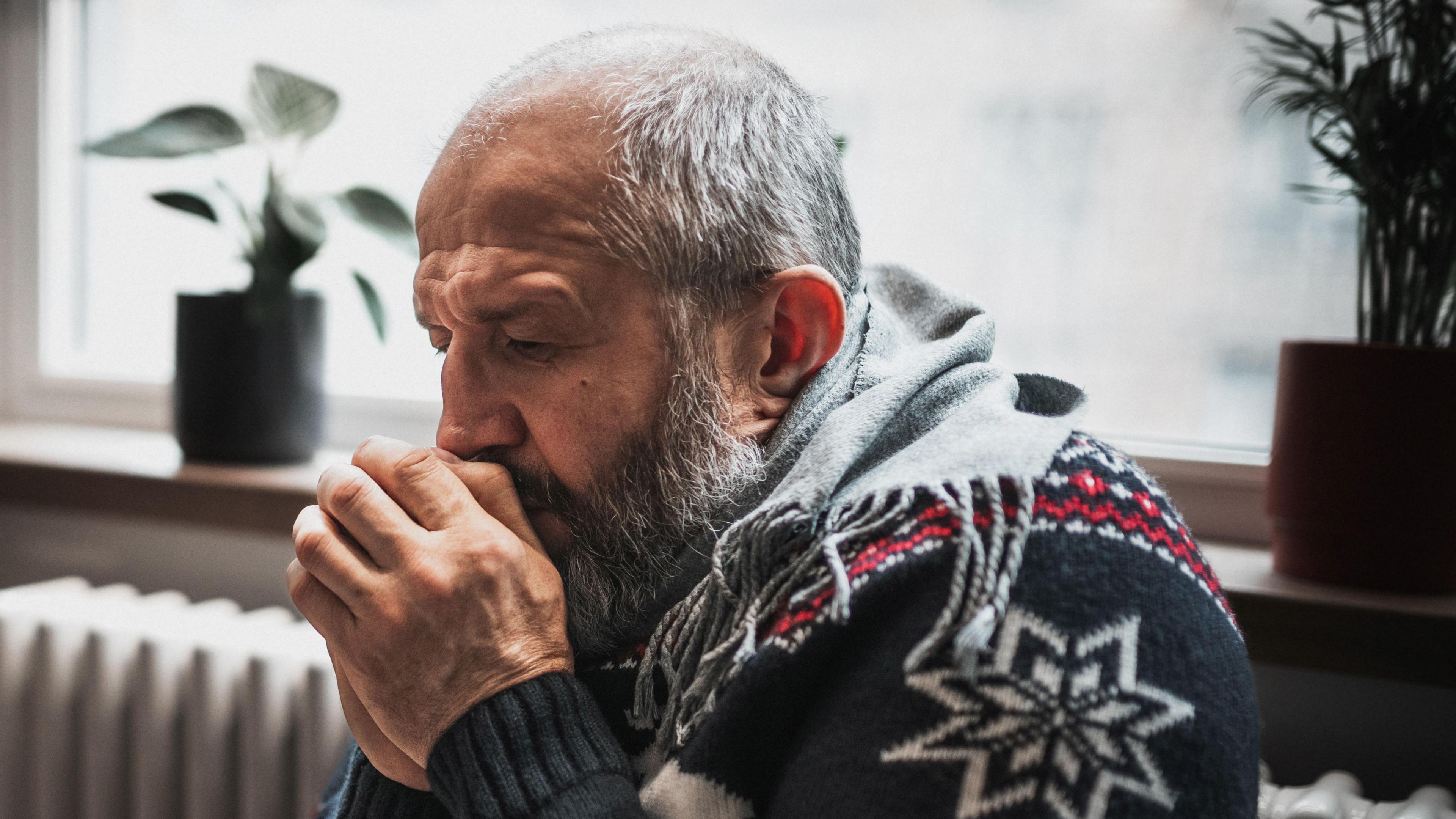 A man in a Christmas jumper huddles for warmth next to a heater