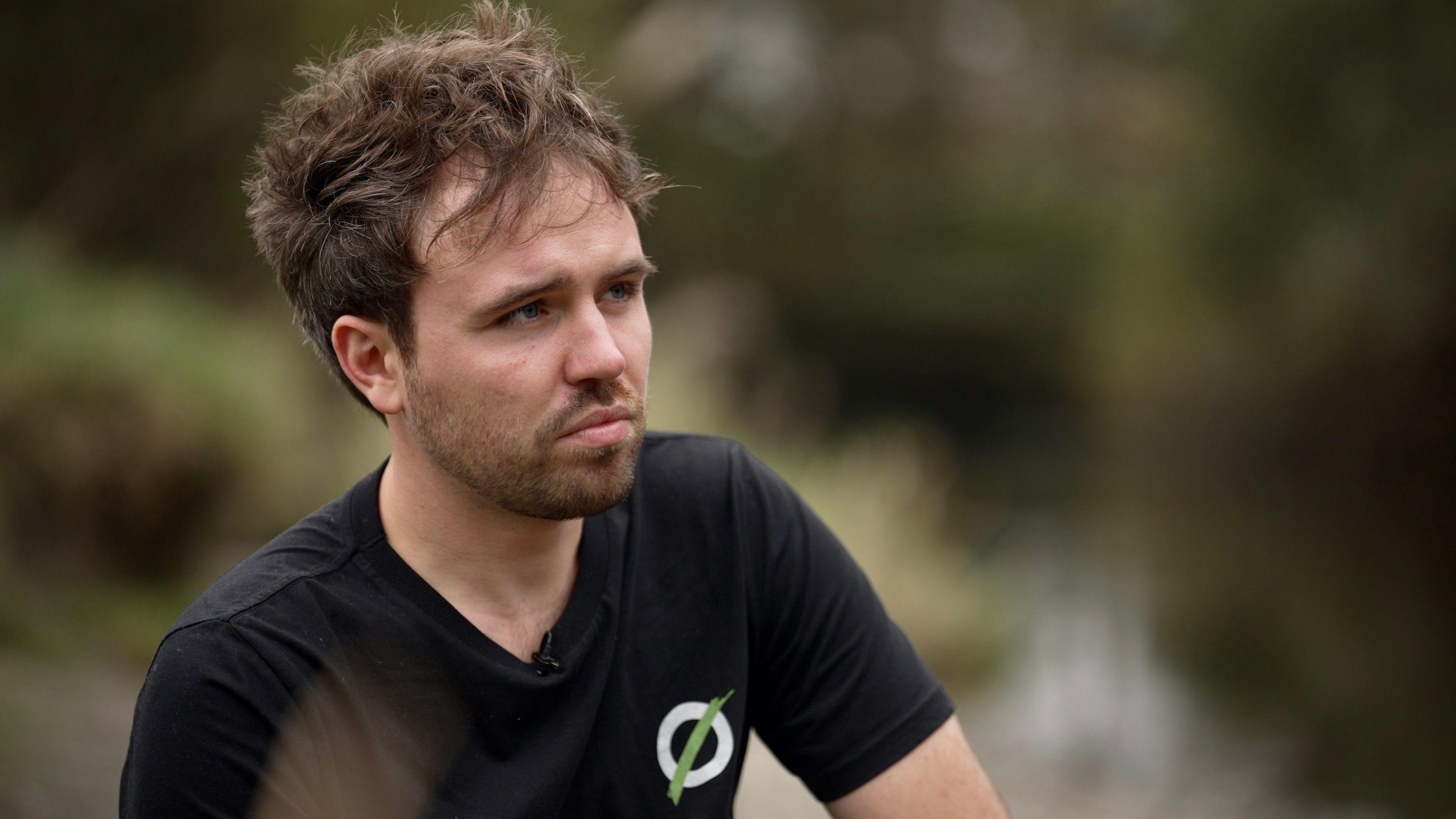 Matt Staniek, wearing a black T-shirt featuring the Save Windermere logo of an O resembling a sewage pipe with a green line slashed across it, pictured by the side of the lake