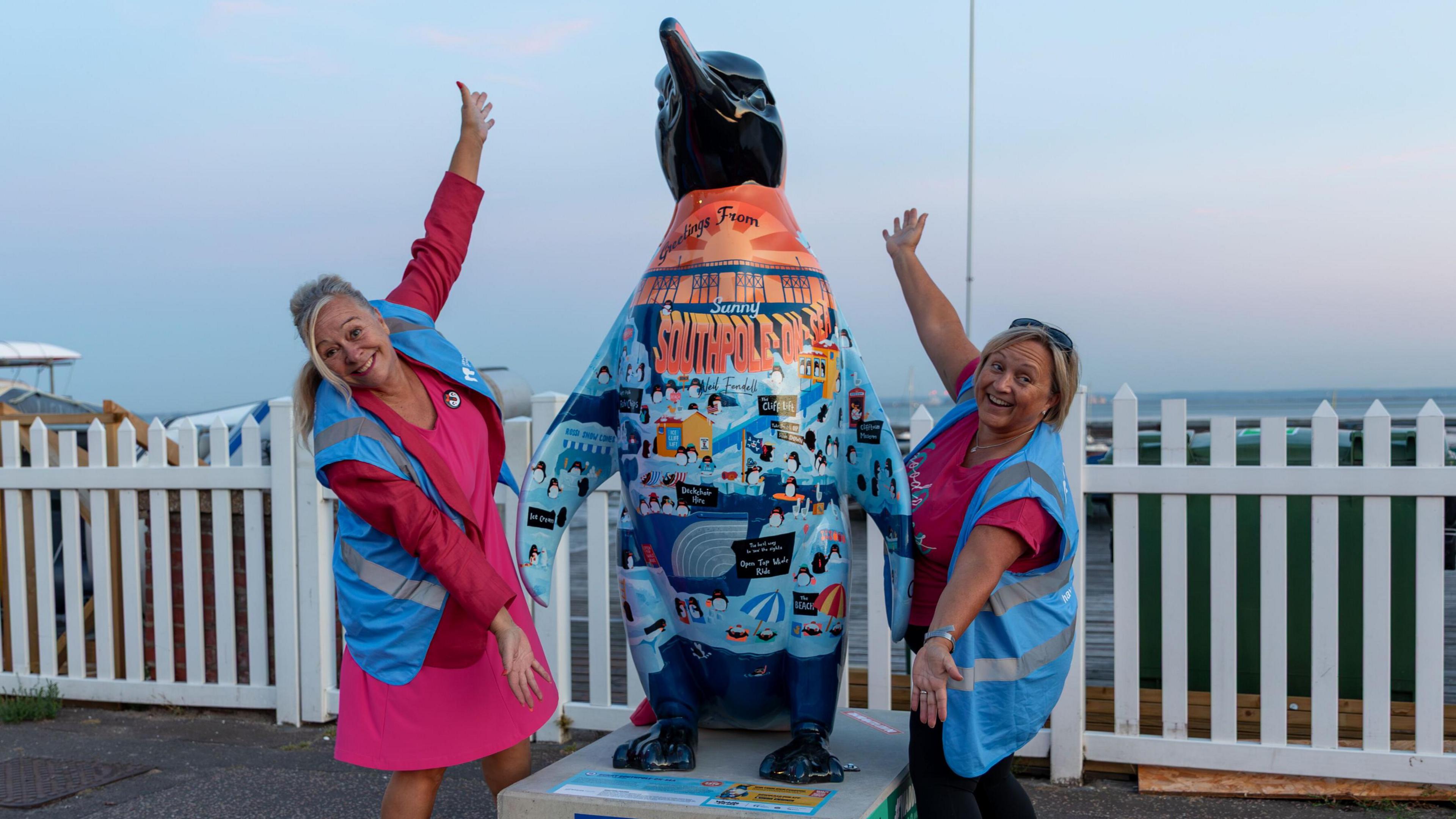 Two people are standing either sides of a penguin sculpture which is in the middle. Both women have blonde hair and are wearing a a blue hi-vis jacket standing in front of a white fence.