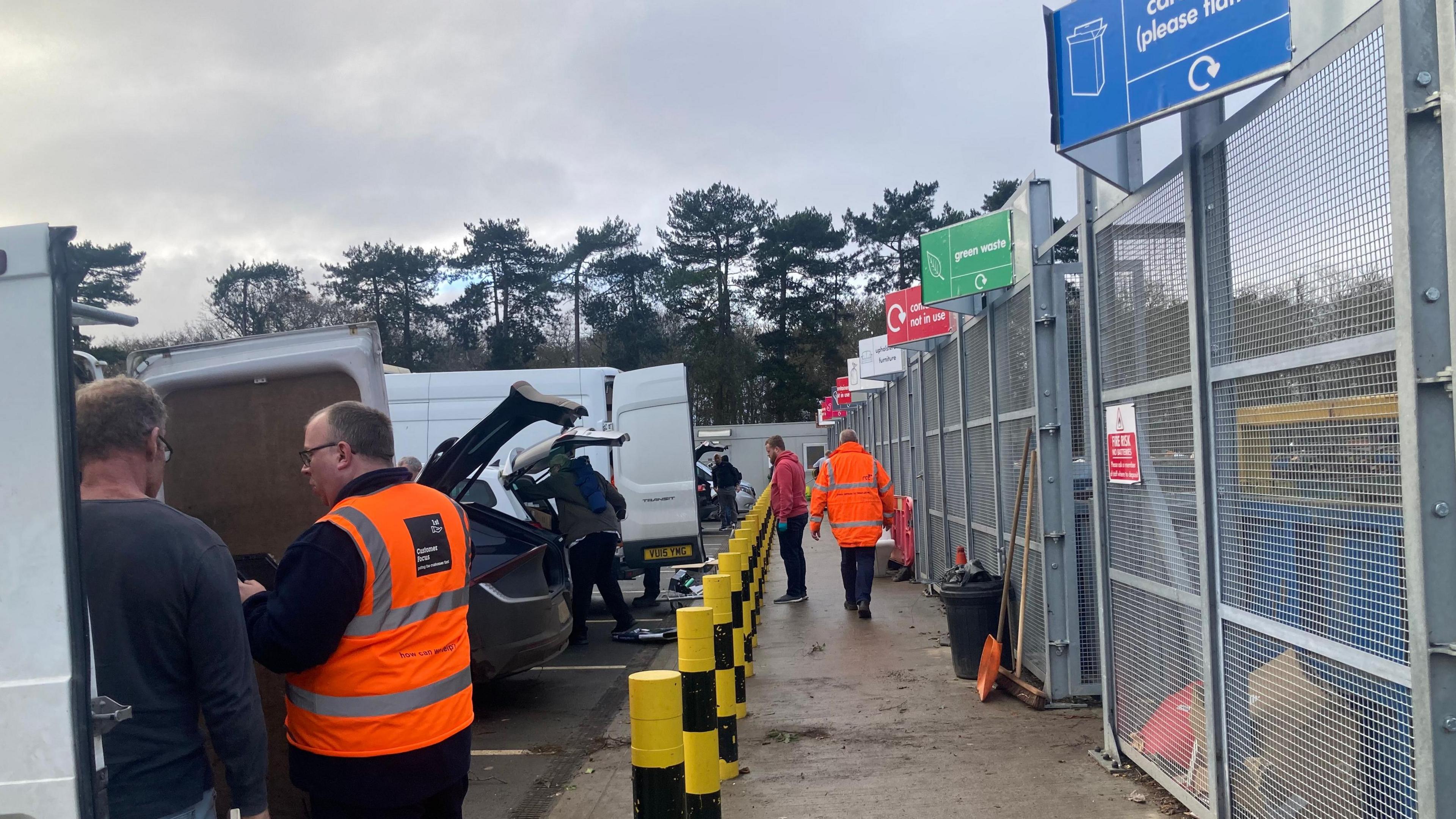 Vehicles are pictured parked up at the entrance of recycling centre's bins. Various areas for different items are marked out by signs. Centre workers in hi-vis jackets can be seen moving around and talking with customers.