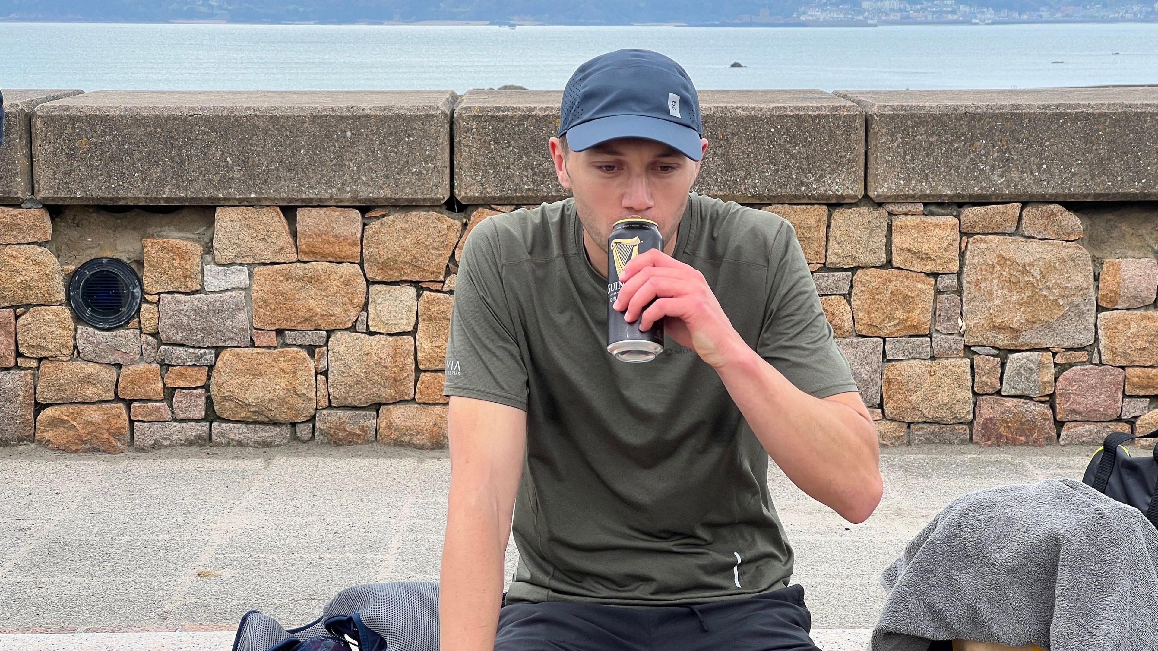 Matt sips from a can of Guinness as he sits on a stone bench. He's wearing a dull green running top and dark shorts and has a grey cap on. Behind him is a stone wall with the sea in the distance behind it.