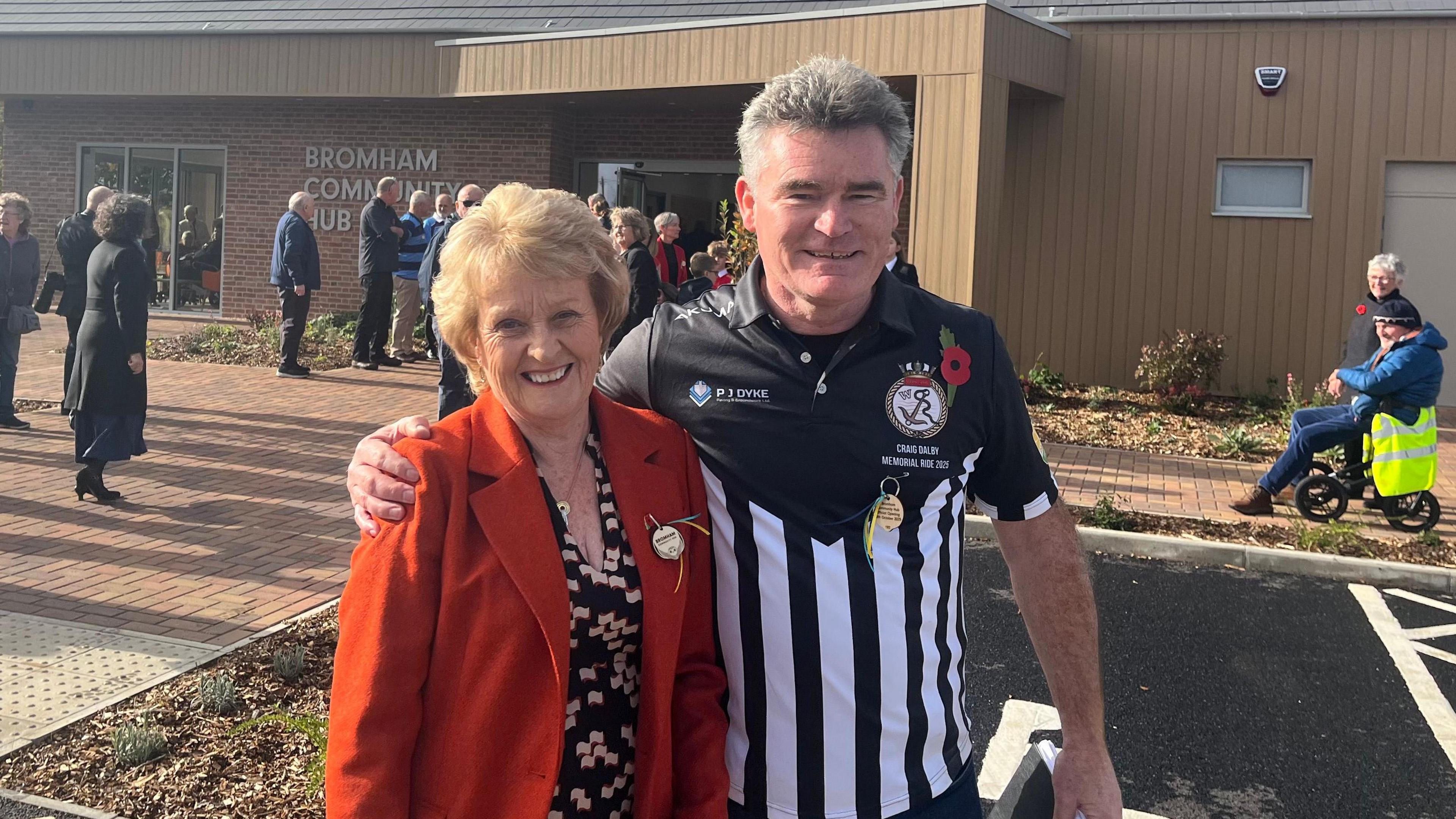 Woman in red jacket and black dress stood next to man in black and white top. In the background, is a wooden building and several people