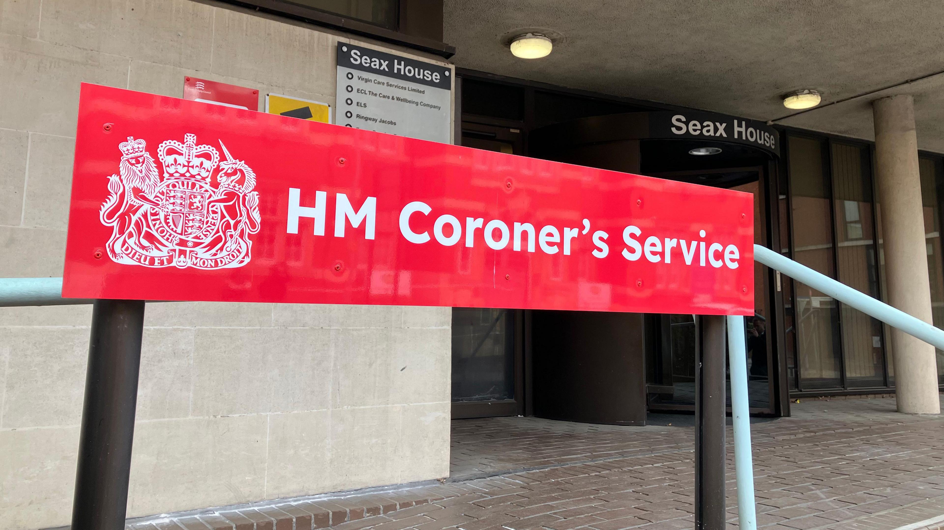 A red sign that says HM Coroner's Service. It is at the entrance to Essex Coroner's Court, which is a cream building that has a black revolving door and brick paving outside.