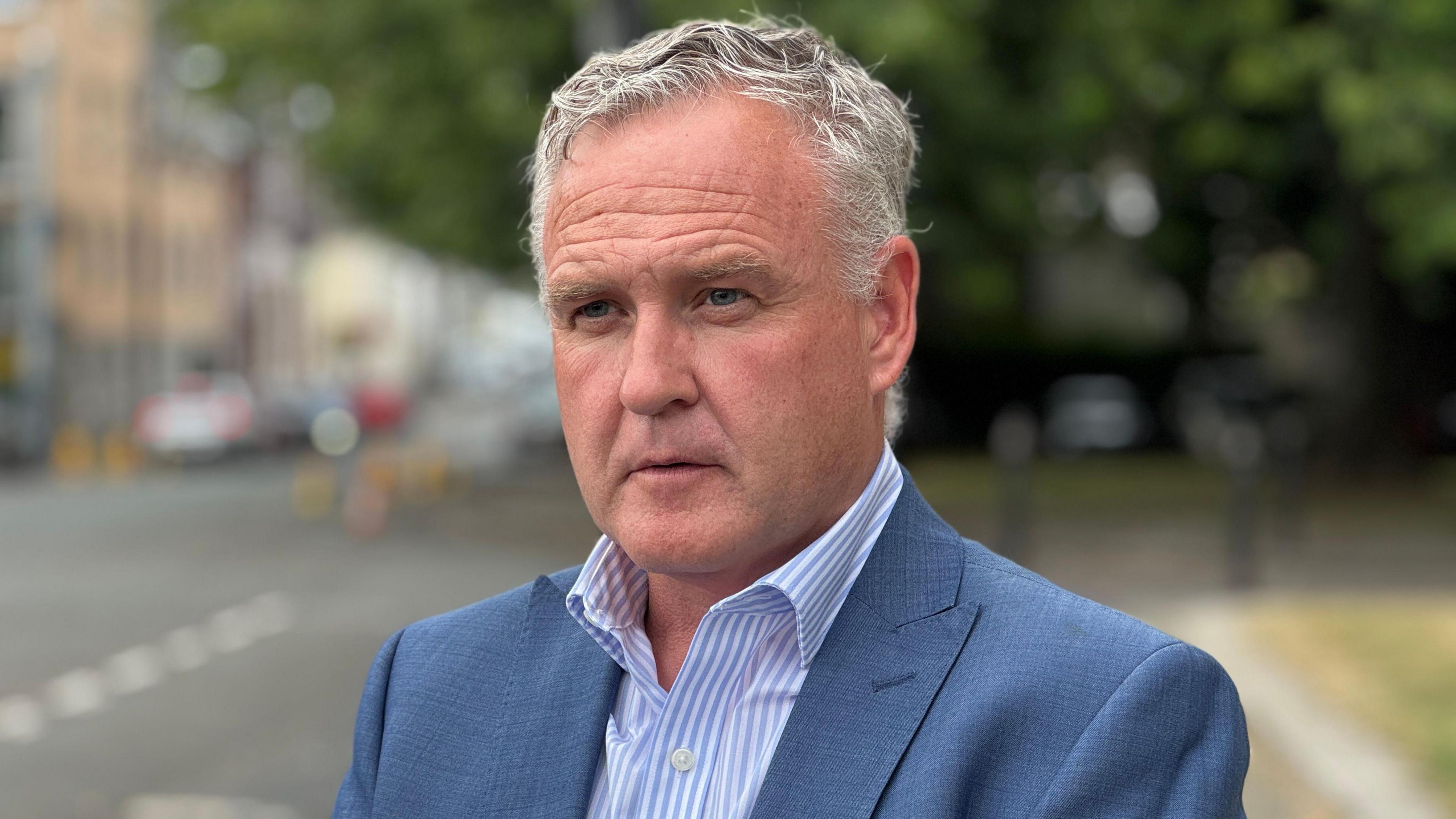 Gethin Edwards in a blue suit and light blue and white striped shirt. He has short grey hair and is standing by a road, which is blurred in the background.