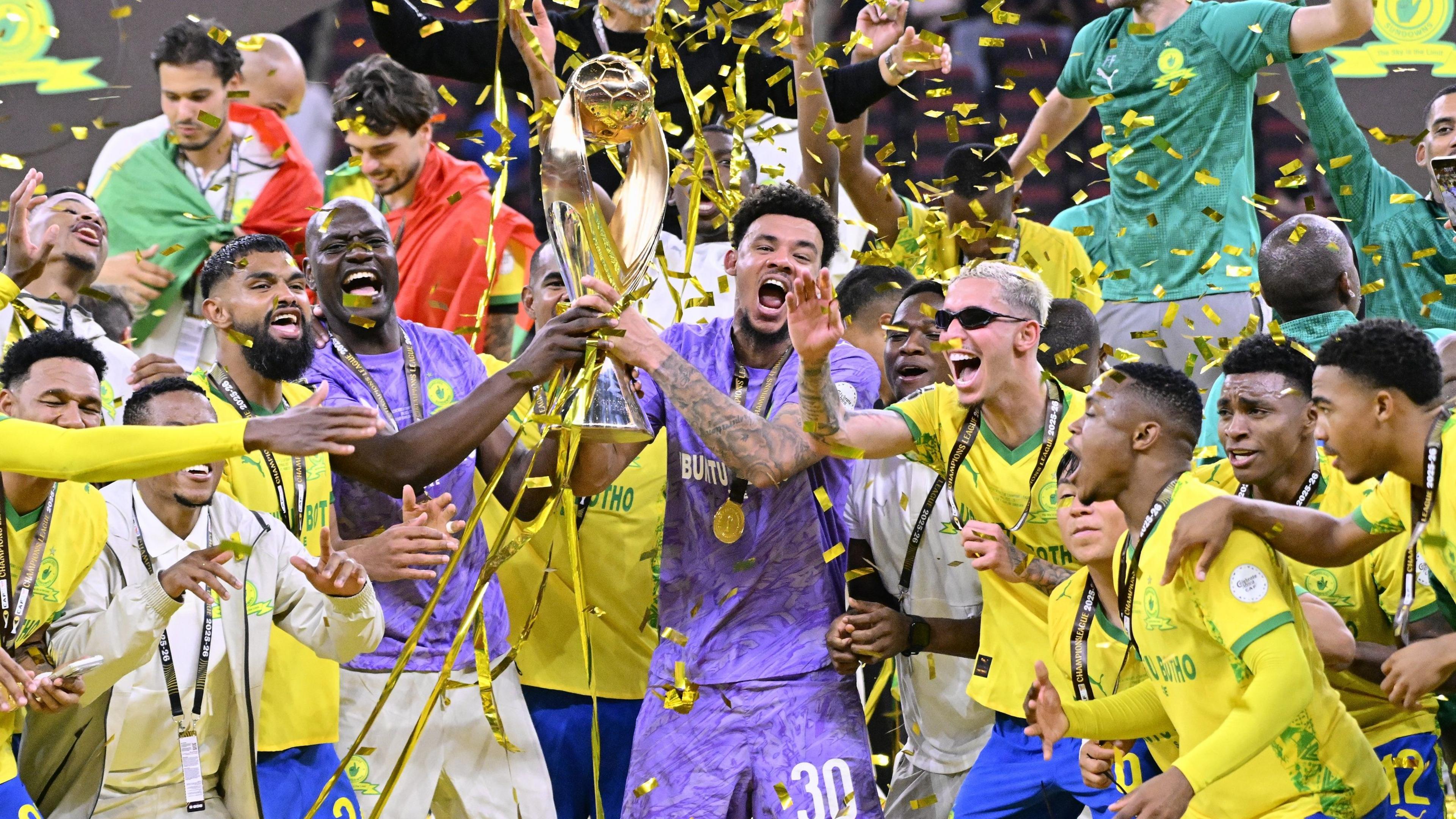 A trophy lift image shows Mamelodi Sundowns players celebrating winning the 2026 African Champions League title, with Ronwen Williams, dressed in purple goalkeeping kit, front and centre lifting the trophy alongside fellow keeper Denis Onyango. The golden trophy consists of two curving sweeps of metal which meet at the top to support a football. Sundowns players in yellow jerseys and blue shorts surround Williams, wearing medals around their necks as golden ticker tape falls around them