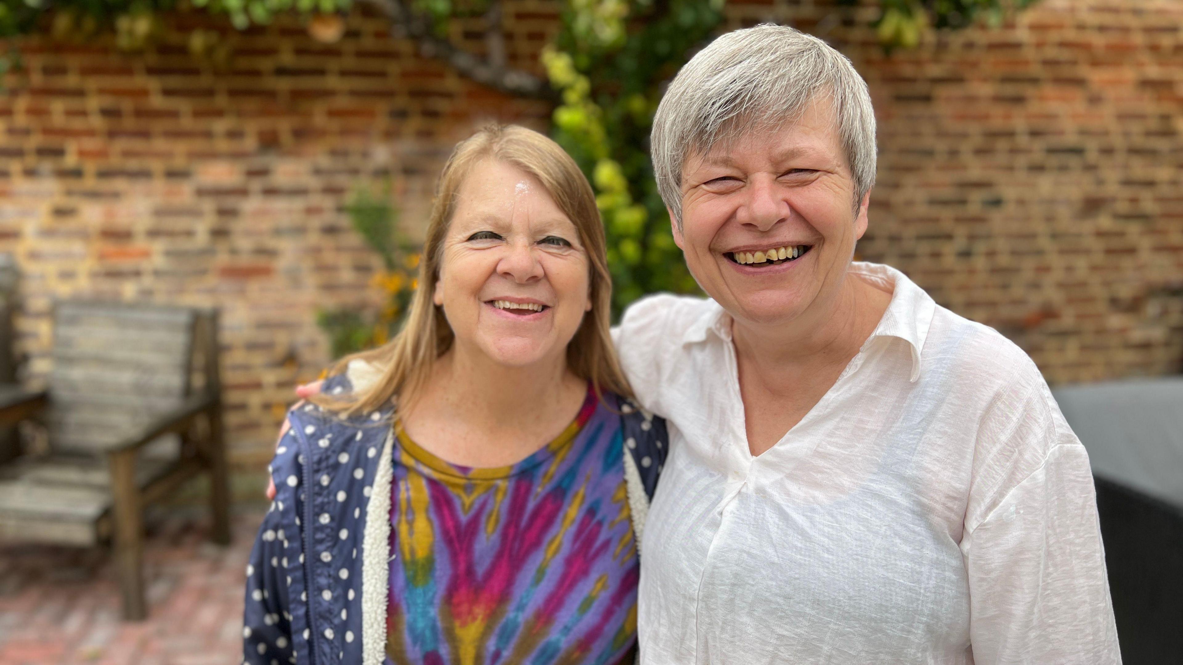 Two female friends smiling with their arms around each other outside in a garden. There is a brick wall and a garden chair behind them.