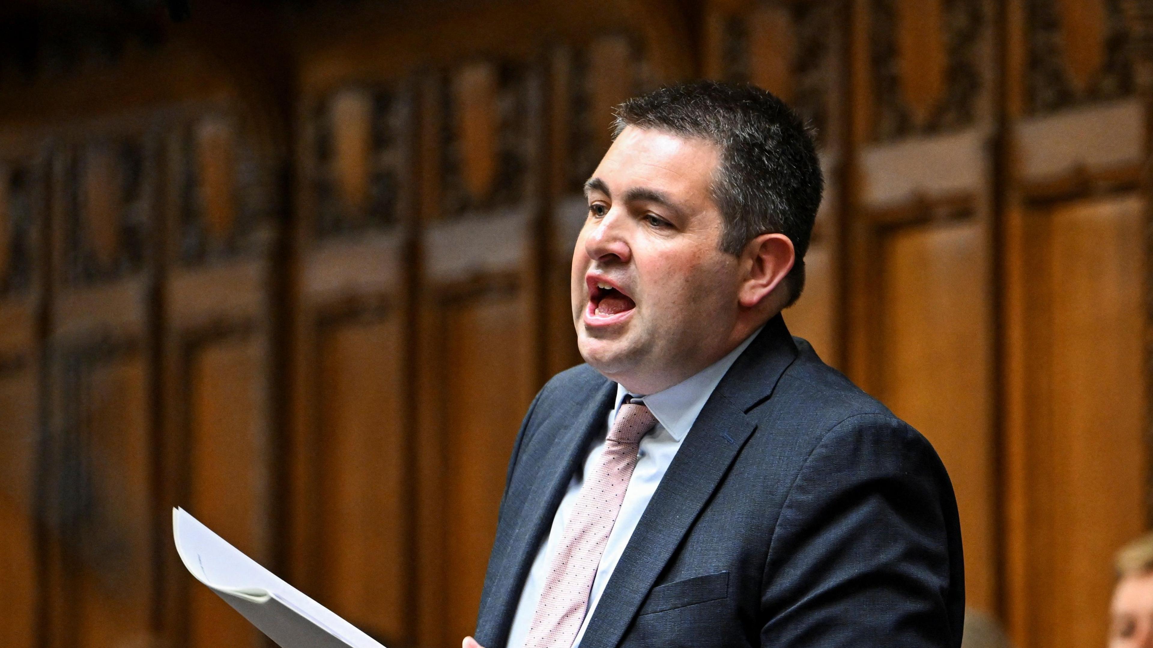 MP Shaun Davies, holding paper and wearing a blue suit, addressing the House of Commons, with wooden panelling behind him